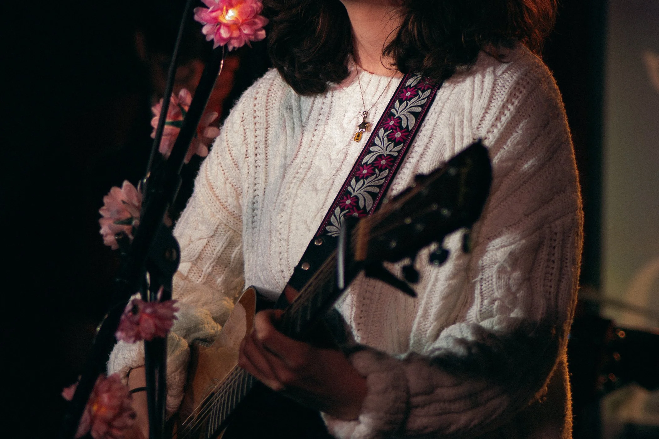 Person wearing a white knitted sweater with floral guitar strap playing an acoustic guitar, with pink flowers on the microphone stand, in a dimly lit setting.