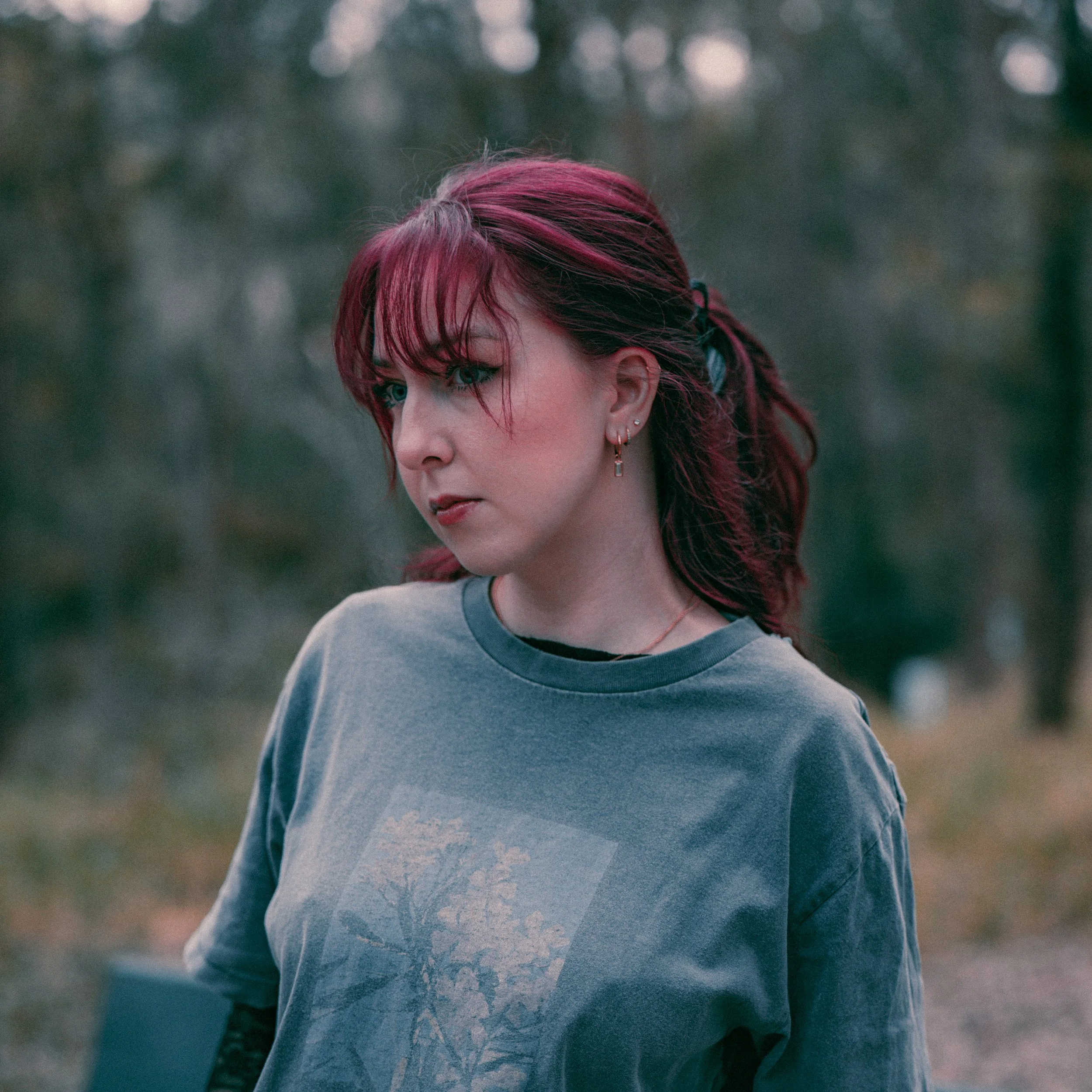 Close-up of a woman's face with red hair and blue eyes, wearing earrings, outdoors in a natural setting.