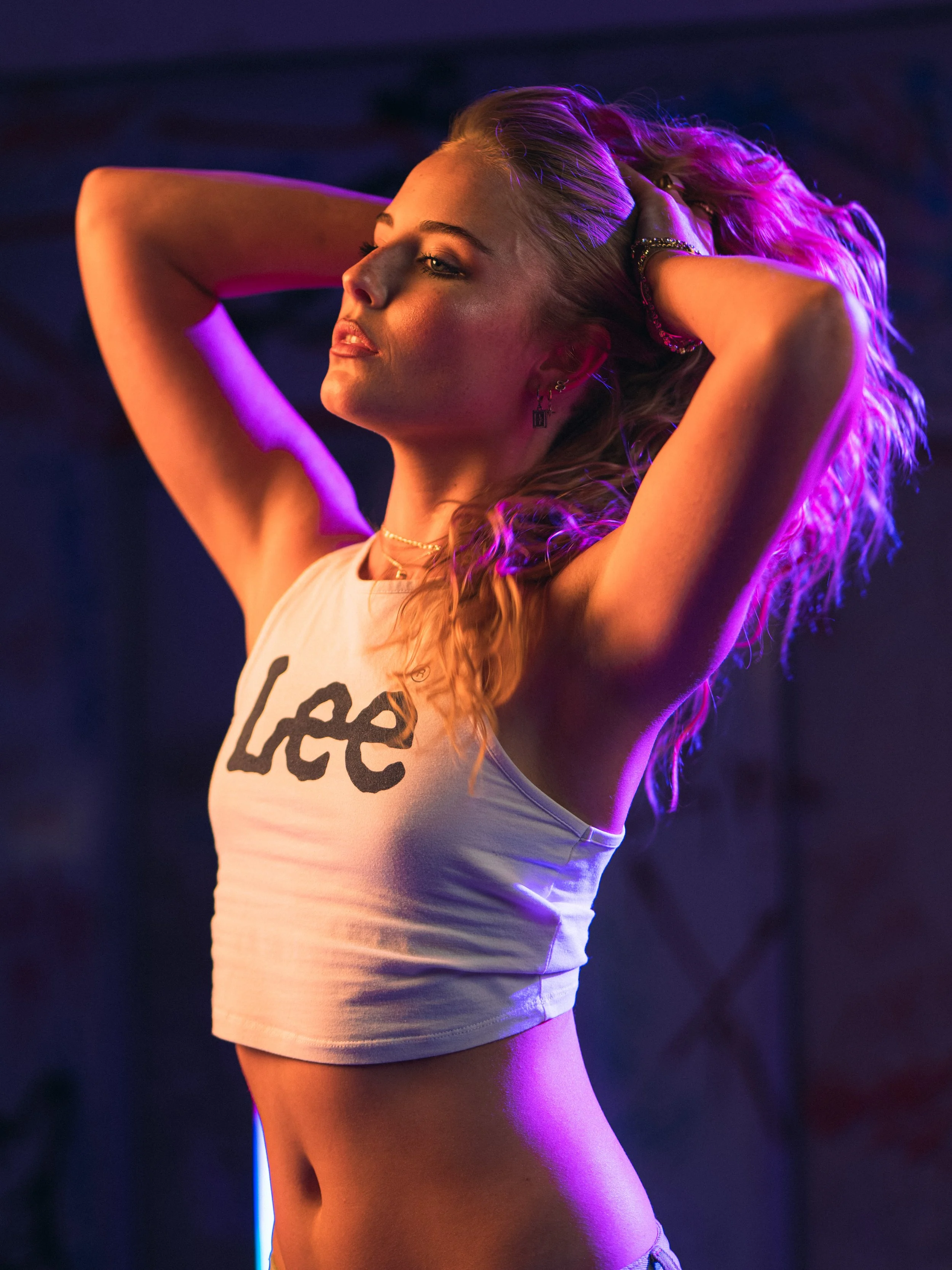 A woman with blonde curly hair posing with her eyes closed, wearing a white tank top with 'Lee' written on it, with purple and pink lighting.