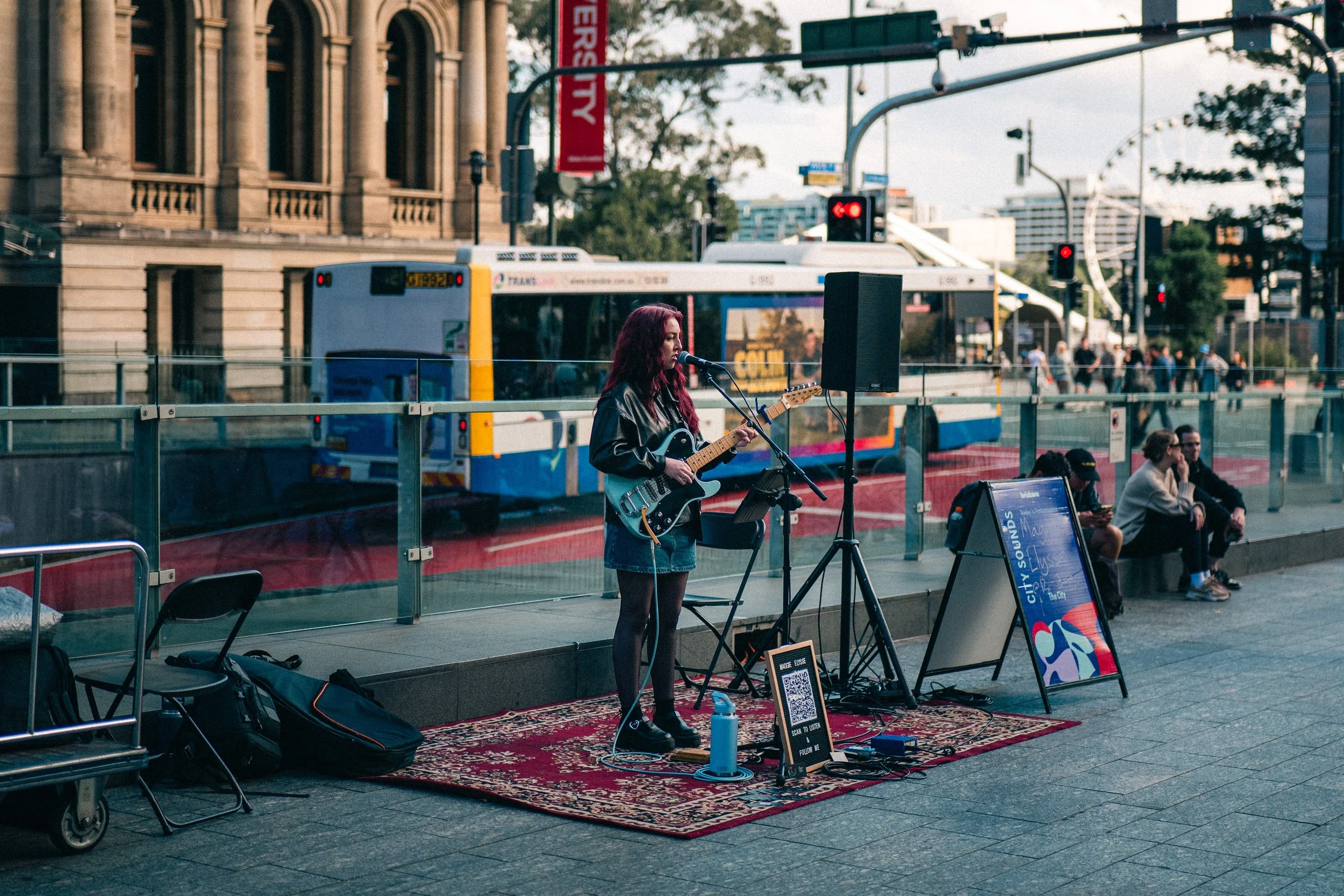 A female musician with red hair performing with an electric guitar at an outdoor city street performance. She is singing into a microphone with a speaker, music stand, and guitar case nearby. Behind her, there is a city bus, a historic building, and 