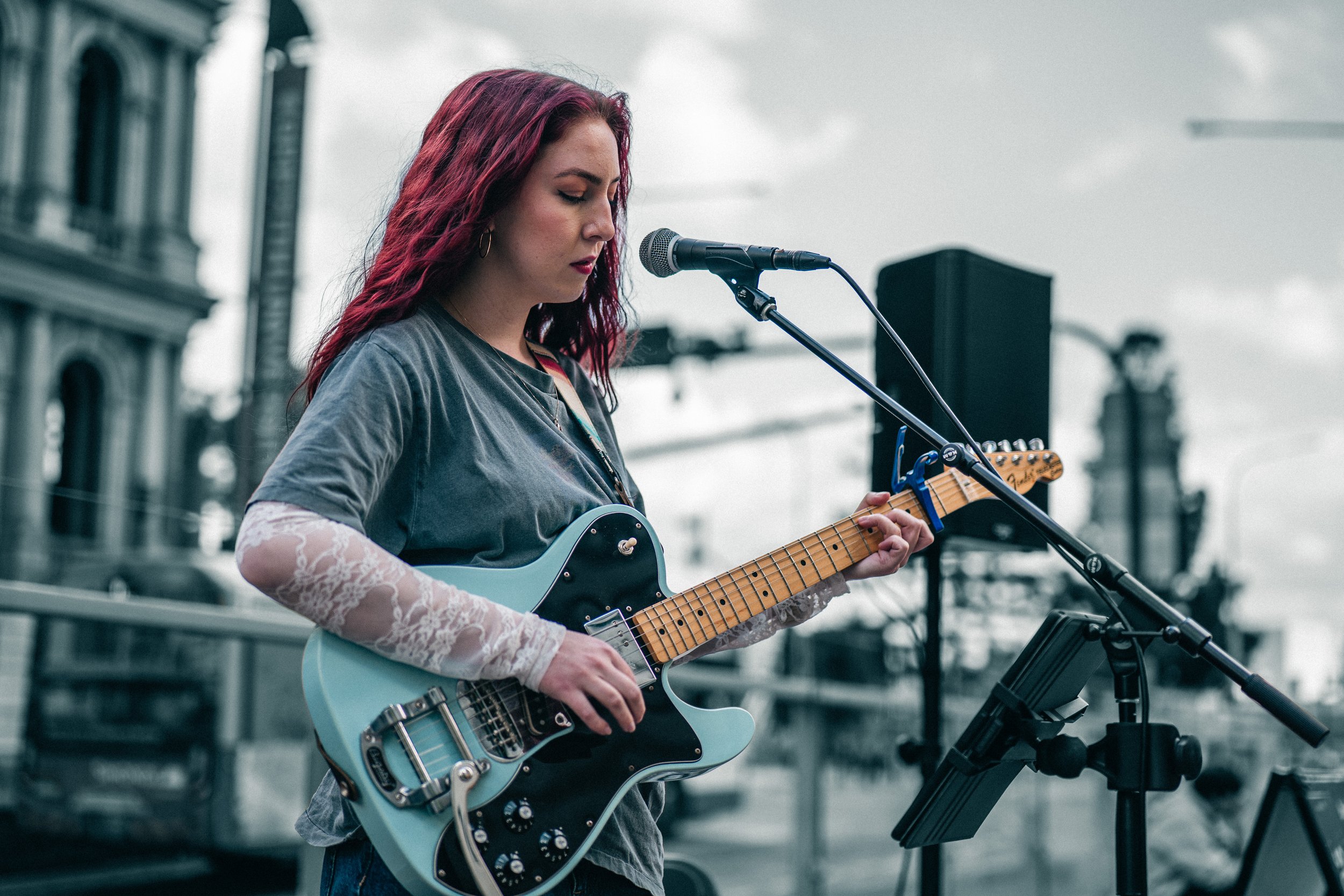 A woman with long red hair playing an electric guitar and singing into a microphone outdoors, with a cloudy sky and city buildings in the background.