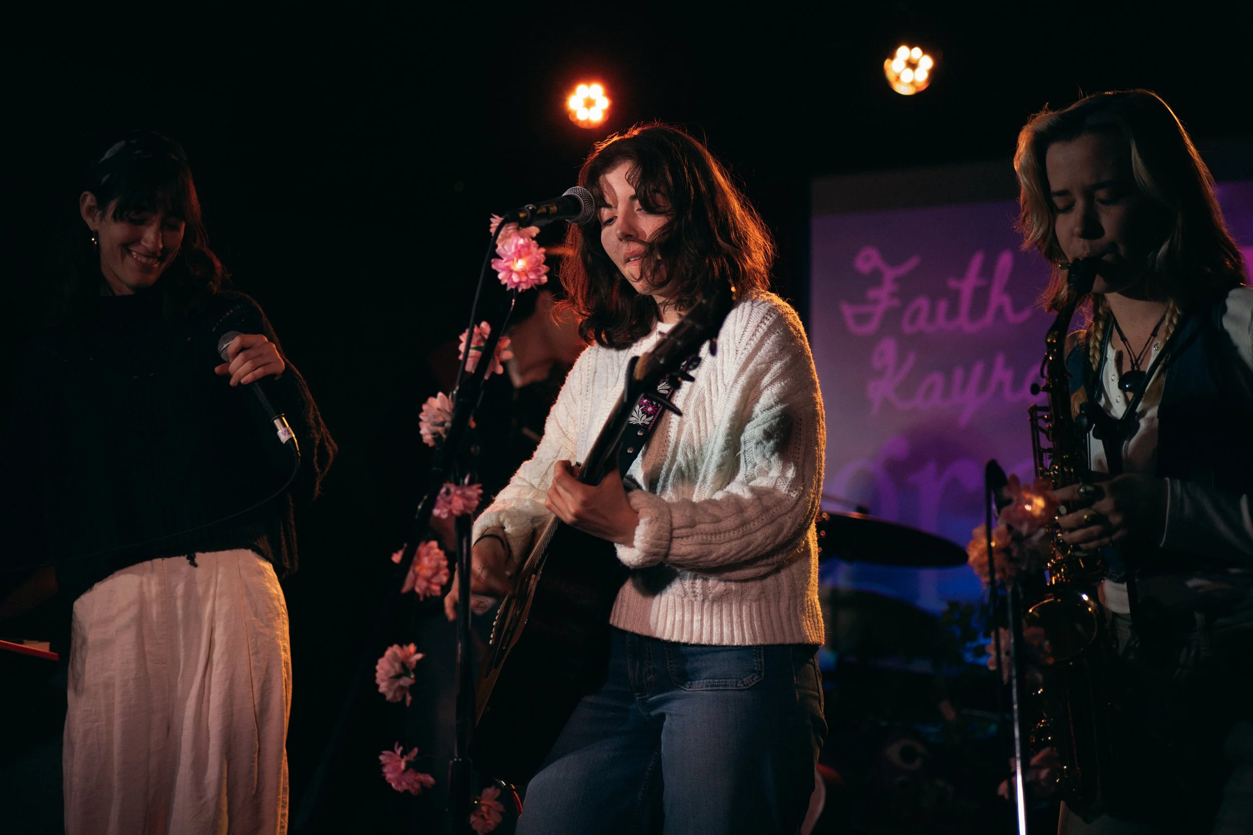 Three women performing music on a dimly lit stage with pink flowers, microphone, guitar, and saxophone, and a purple-lit sign that says 'Faith & Kathleen' in the background.