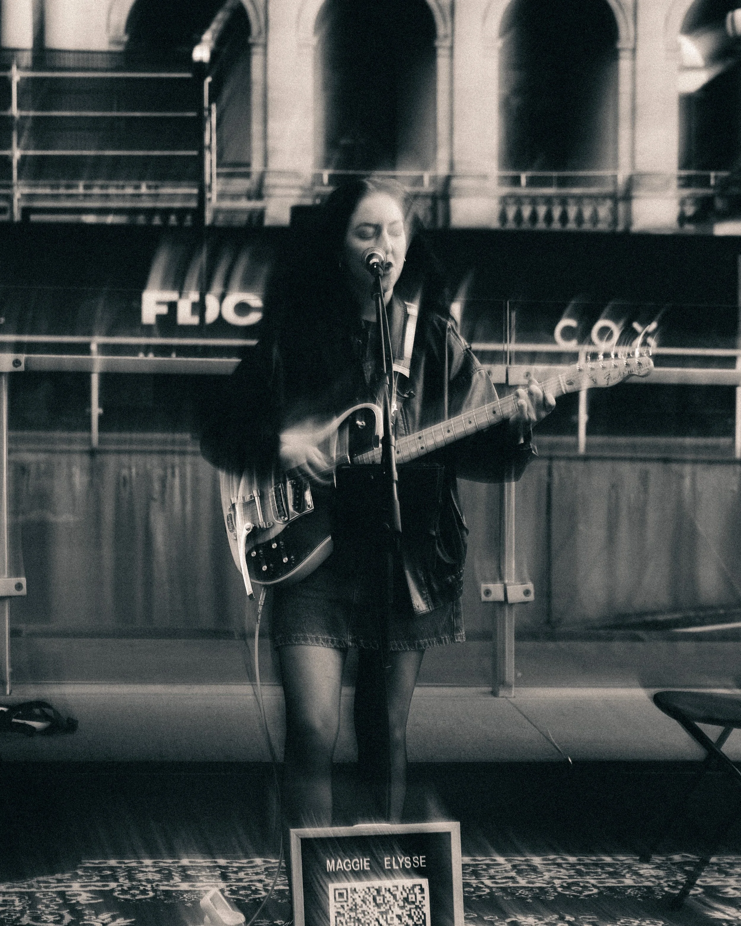 Black and white photo of a woman singing into a microphone while playing an acoustic guitar, standing in front of a location marker with the name Maggie Elyse and a QR code.