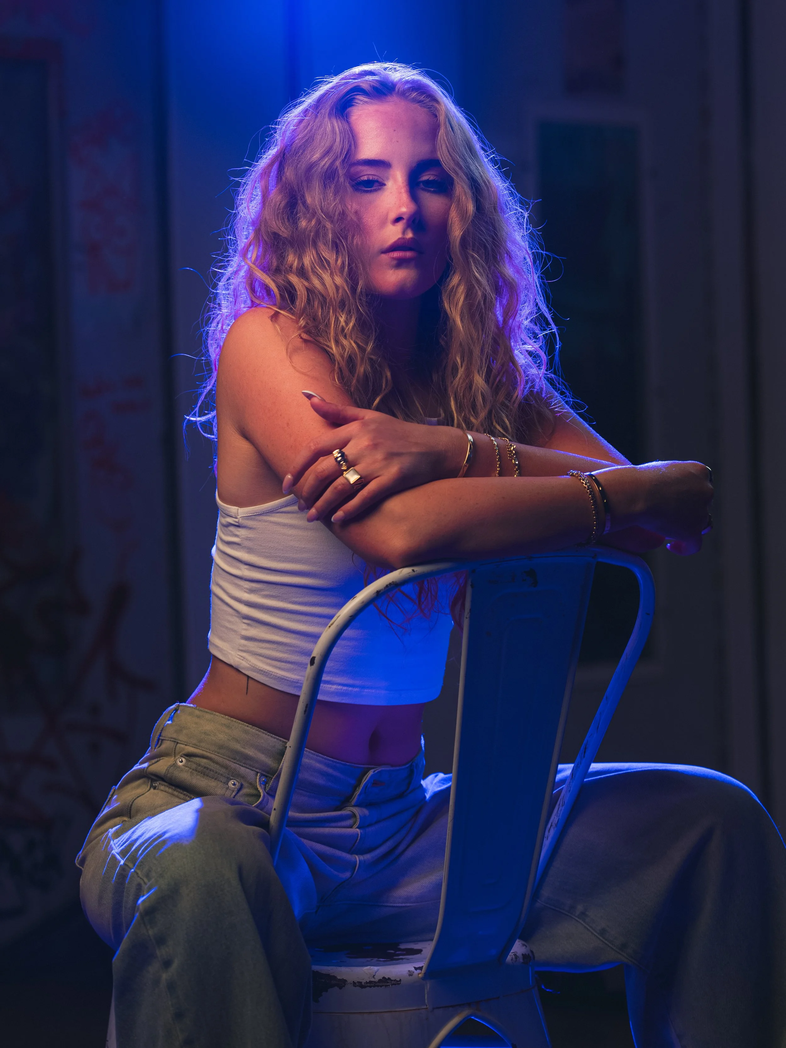 A woman with long curly blonde hair sitting on a white chair, wearing a white tank top and beige pants, with her arms crossed and looking at the camera in a dark room with blue lighting.