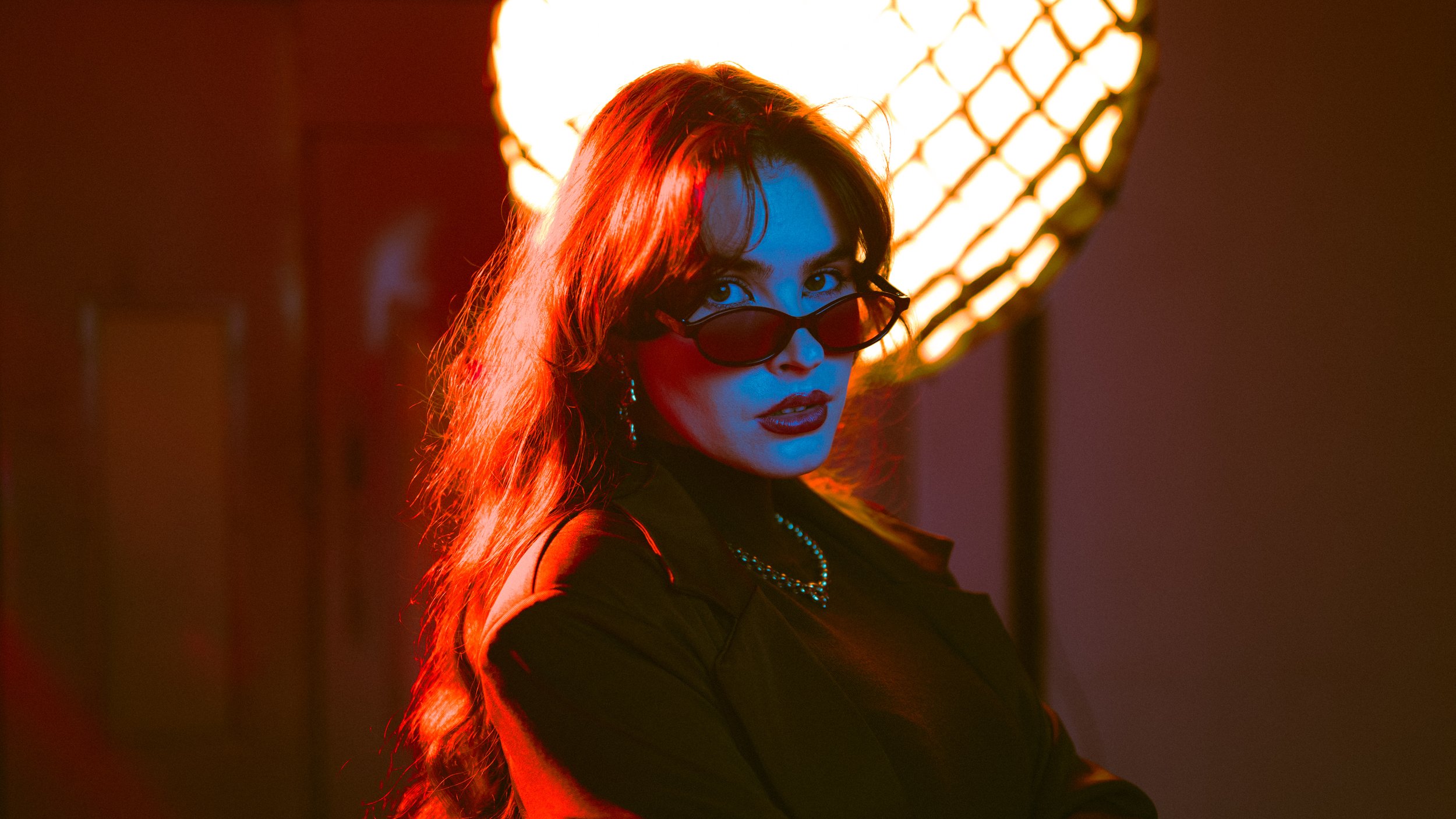 A woman with long hair and sunglasses posing in front of a bright, warm-colored light.