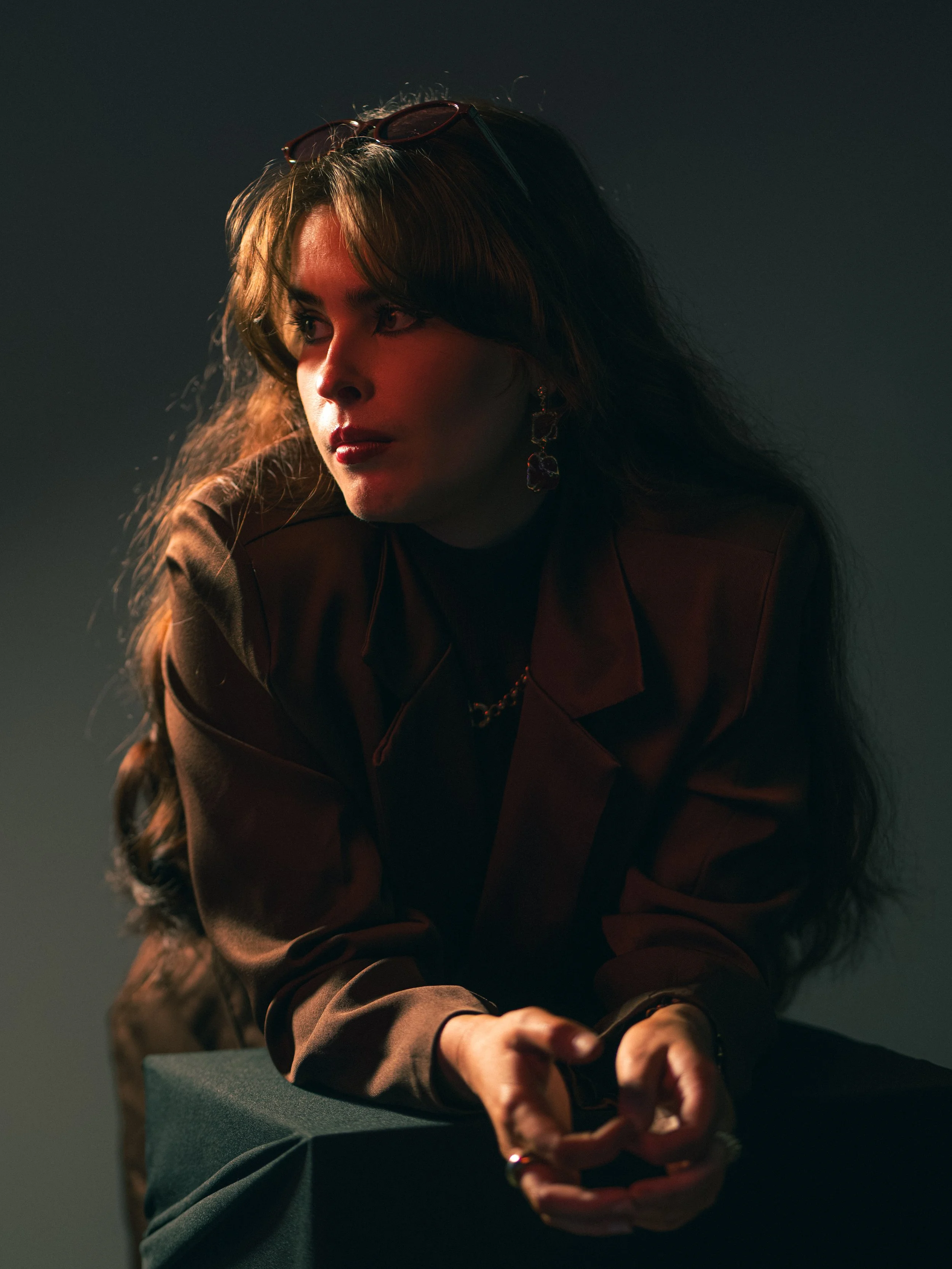 A woman with wavy brown hair, wearing sunglasses on her head, jewelry including earrings and a necklace, is leaning forward with her hands clasped in front of her. The background is dark, with lighting highlighting her face and upper body.