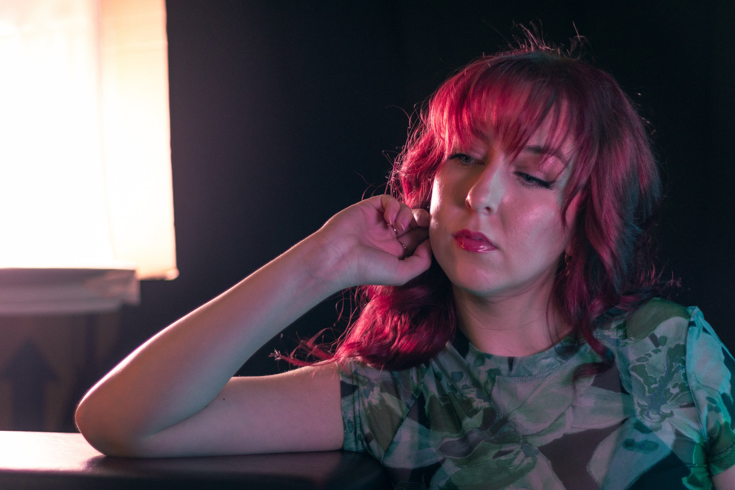 A woman with wavy pink hair, wearing a camouflage shirt, sitting at a desk with her hand near her ear, in a dimly lit room with a bright light source on the left side.