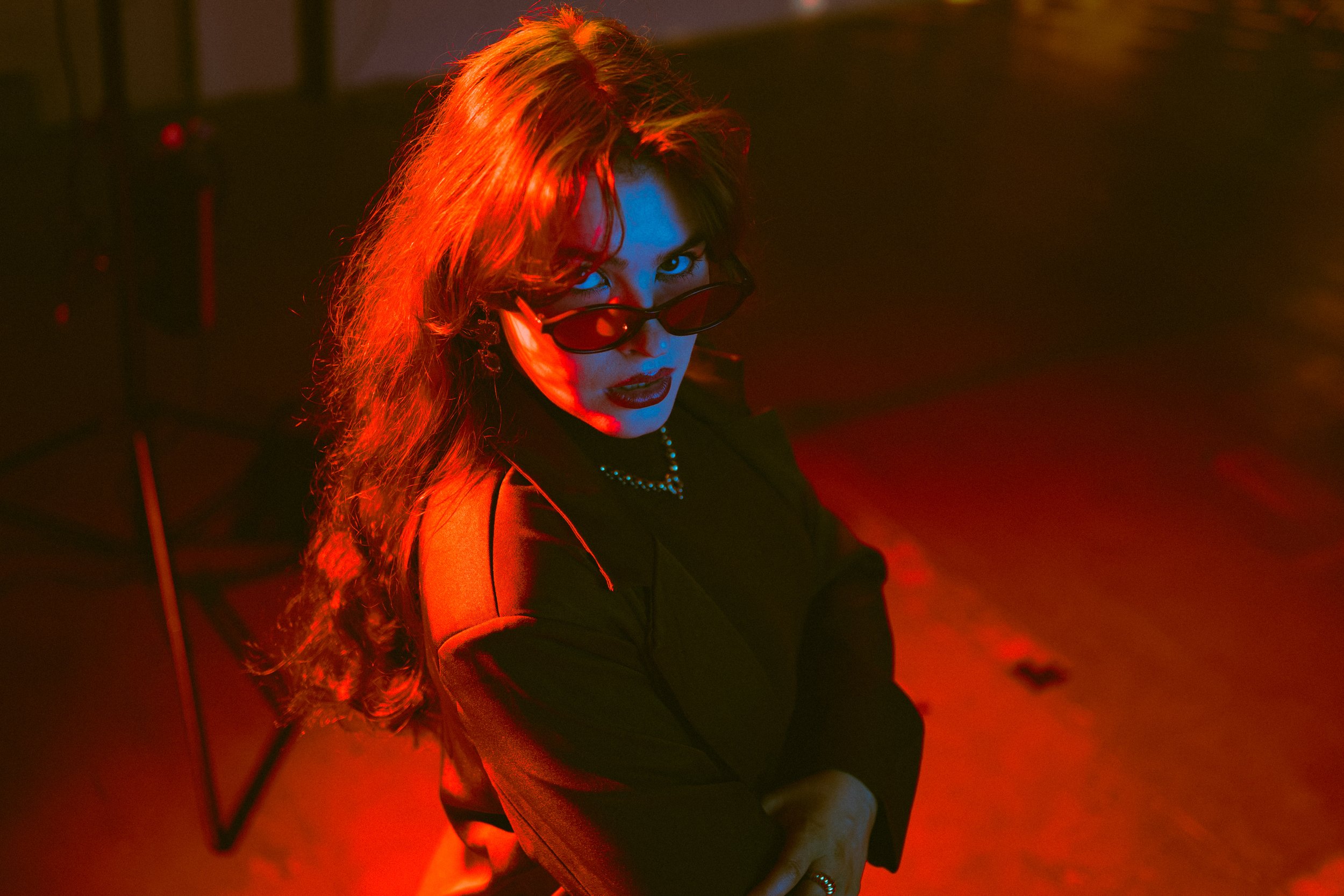 A woman with long, curly hair and sunglasses is looking at the camera in a dimly lit room with red and blue lighting.
