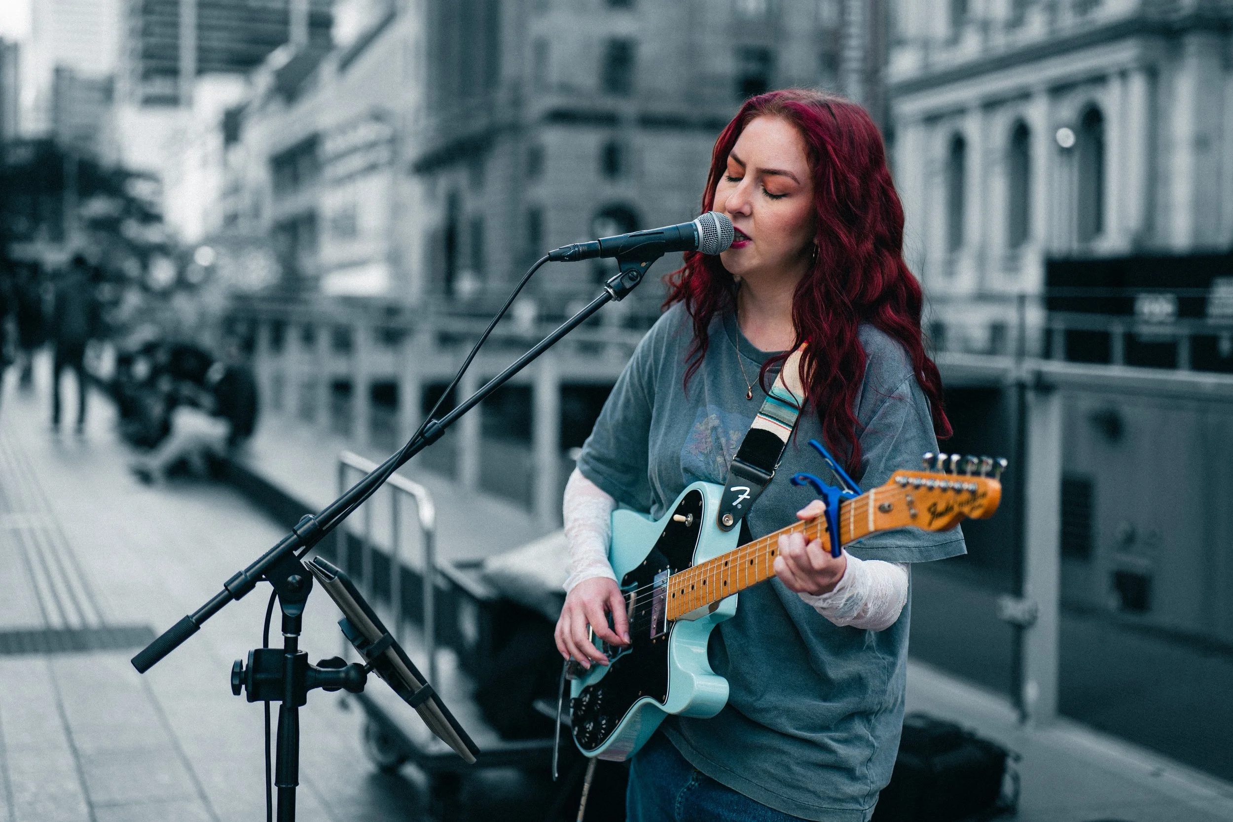 A woman with red hair playing an electric guitar and singing into a microphone outdoors in an urban area.