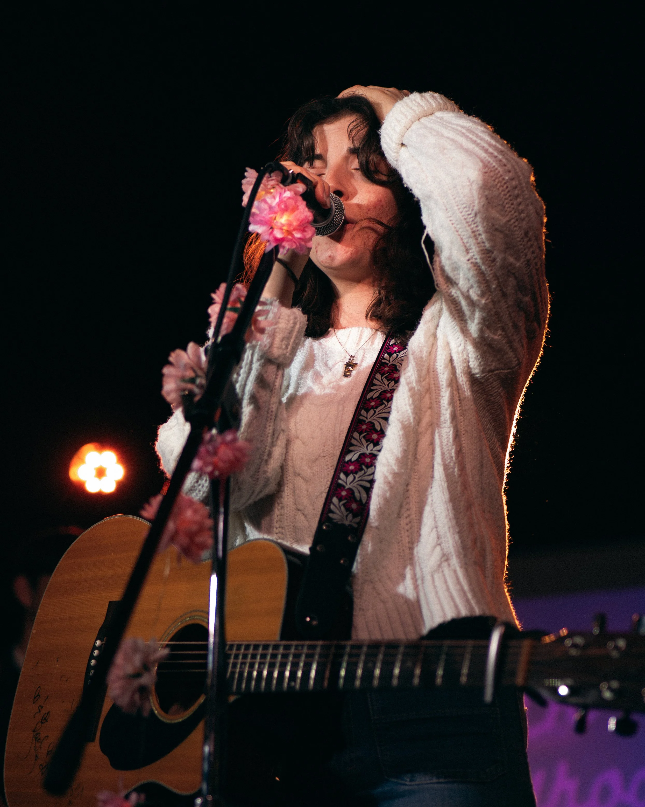 Young woman singing into a microphone decorated with pink flowers while playing an acoustic guitar, wearing a white knitted sweater against a dark background with stage lights.