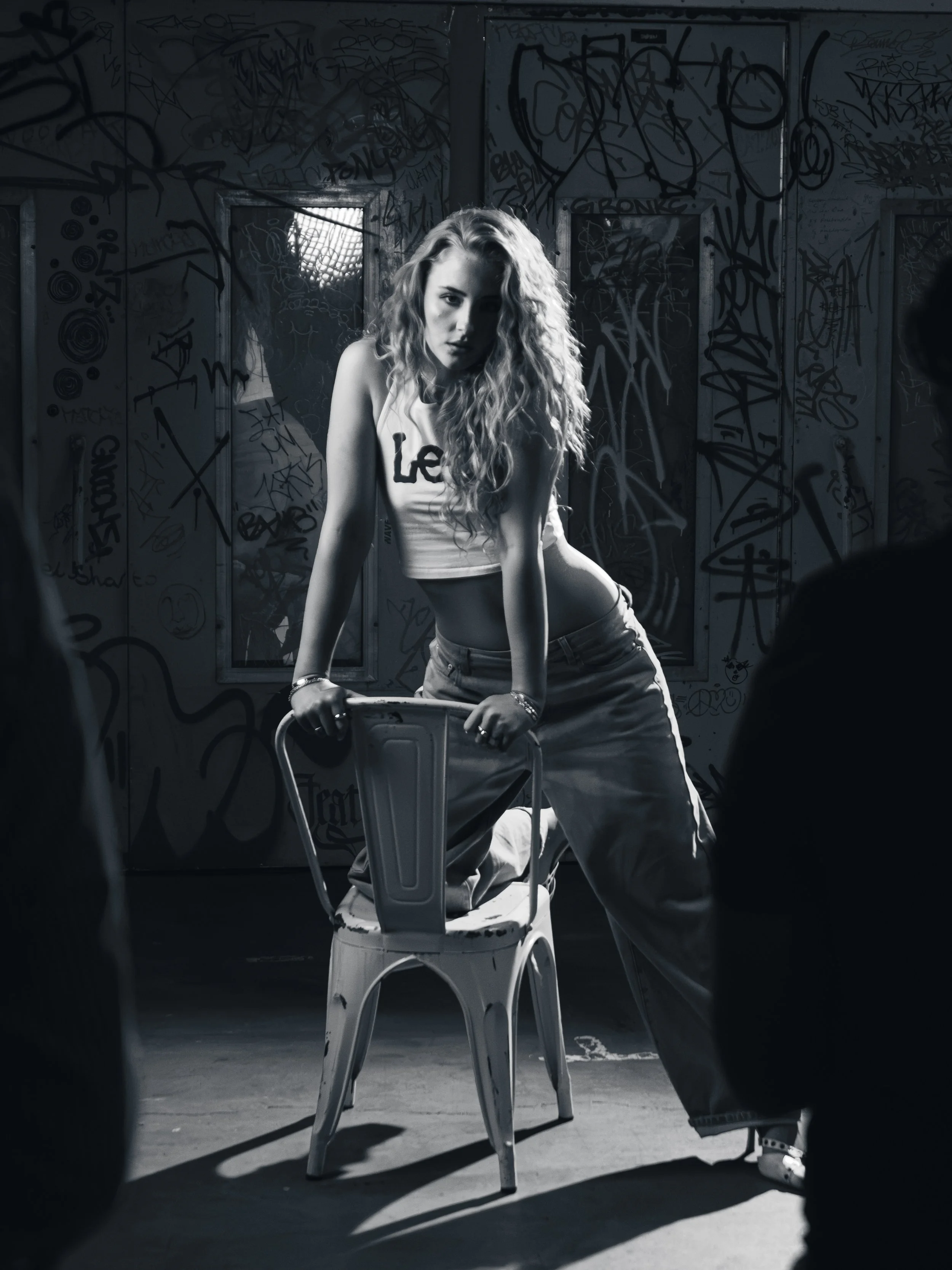 Black and white photo of a woman with curly hair leaning forward on a chair, in a graffiti-covered room with windows in the background.