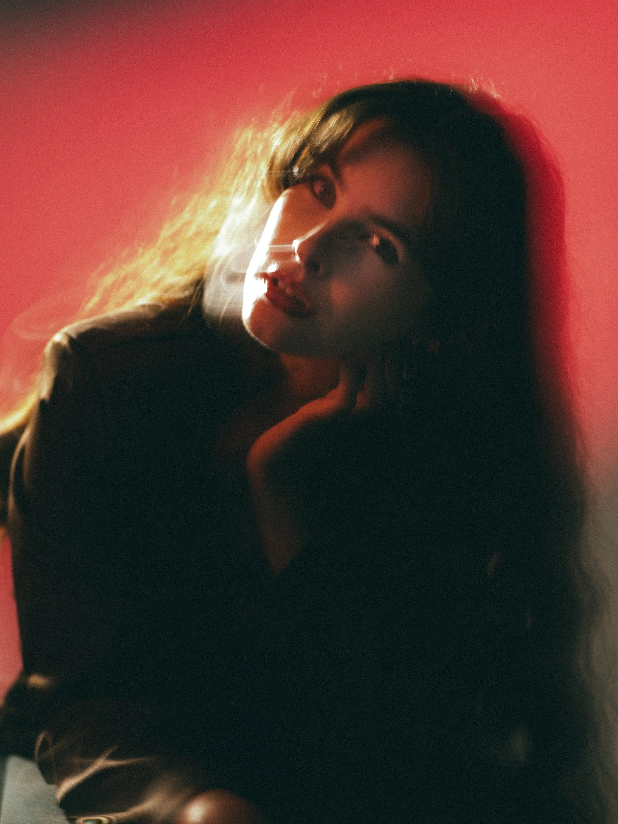 A woman with long hair looking slightly to the right, her face illuminated with soft lighting against a reddish background.