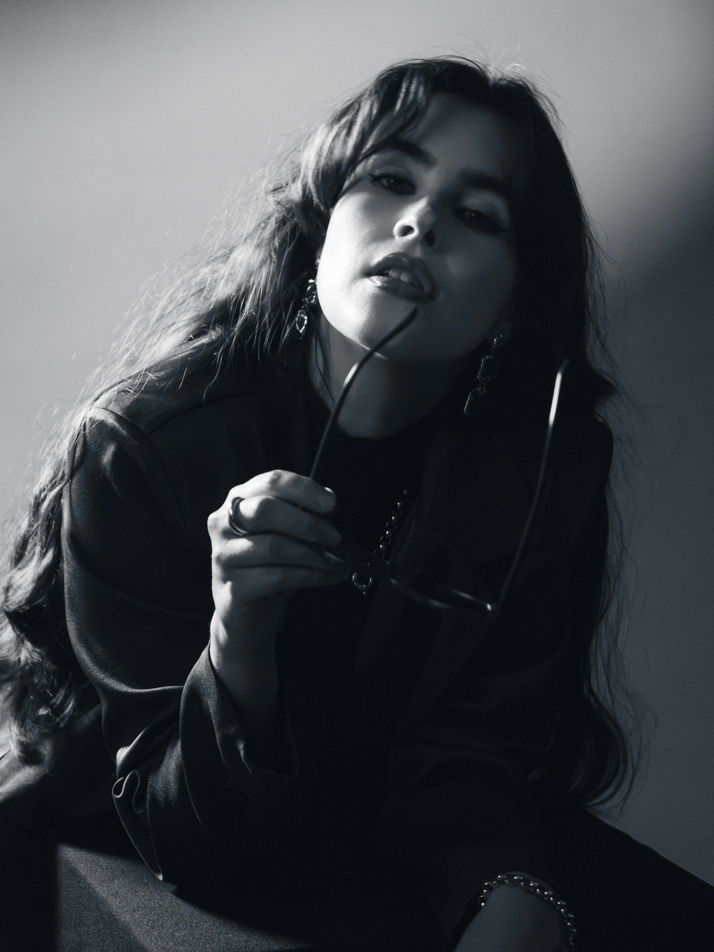 Black and white photo of a woman with long, wavy hair, wearing earrings, holding glasses, looking at the camera with a confident expression.