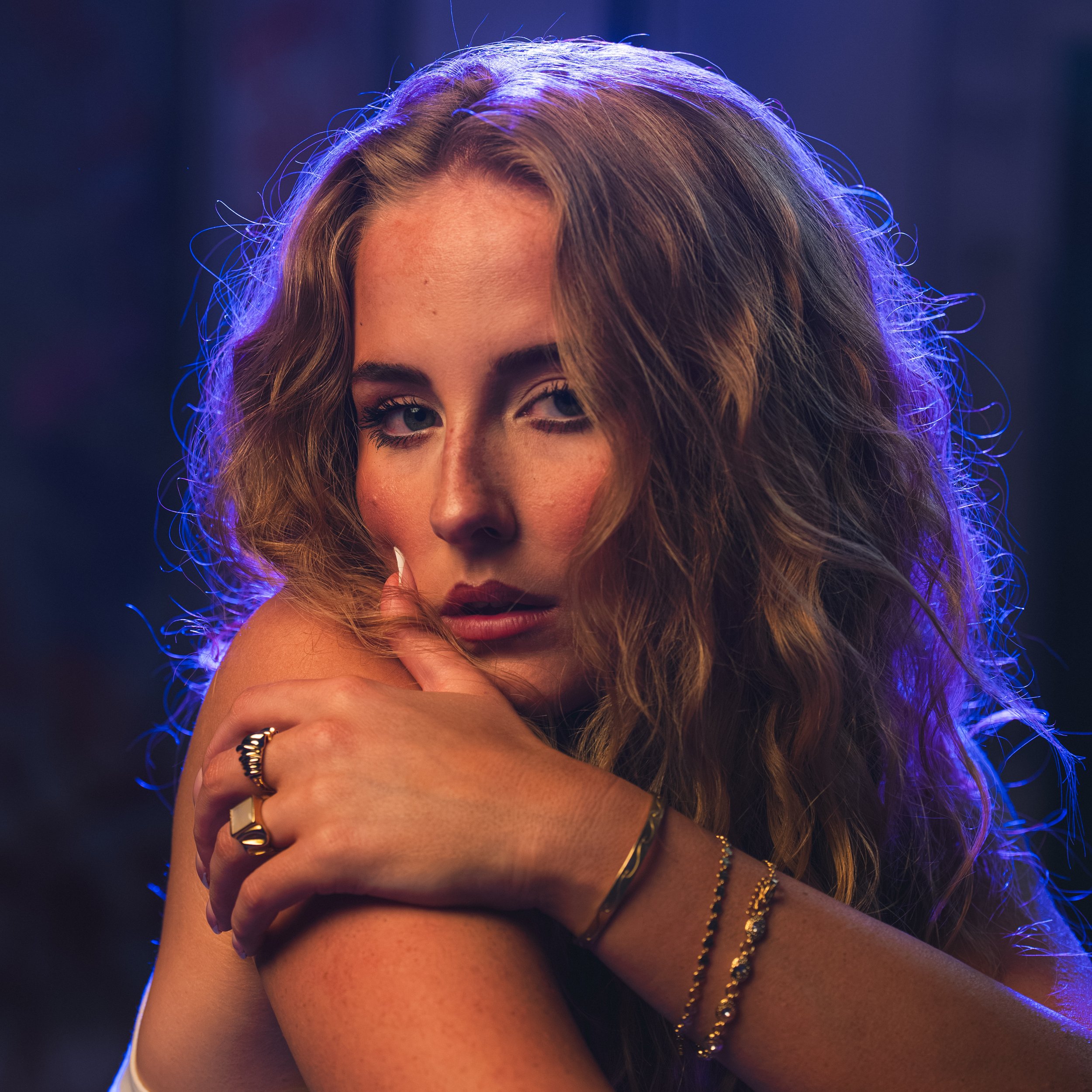 A woman with wavy hair partially covering her face, wearing multiple gold necklaces, her hand with rings near her chin, illuminated by blue-purple lighting in a dark background.