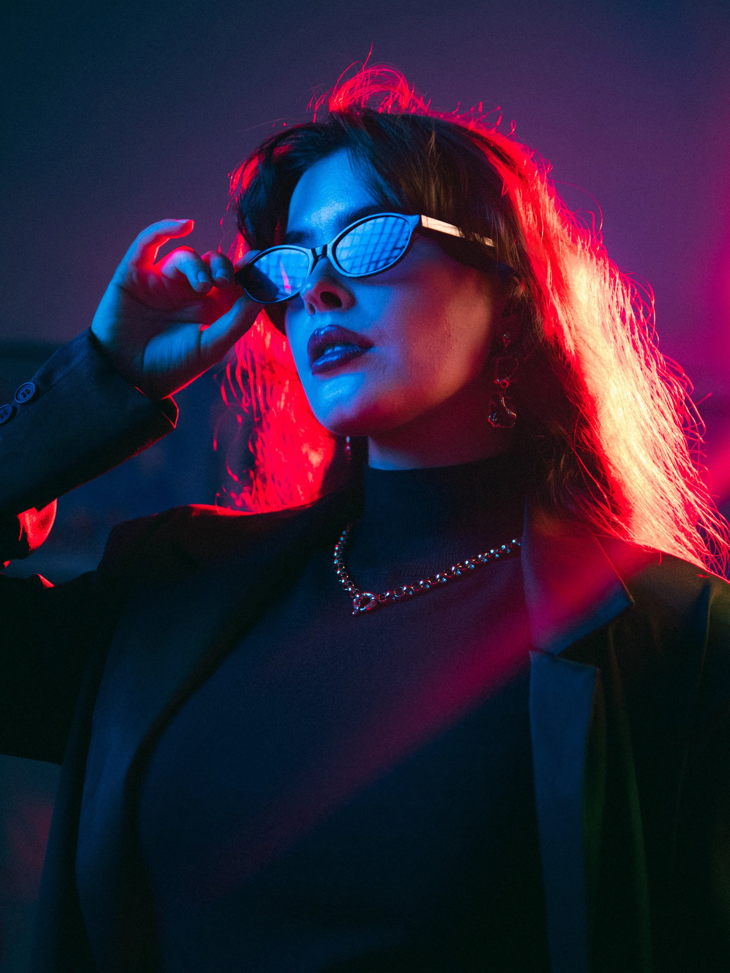 A woman with dark hair and dark lipstick wearing sunglasses with a grid pattern, illuminated by blue and red lighting, dressed in black, with a chain necklace, posing against a dark background.