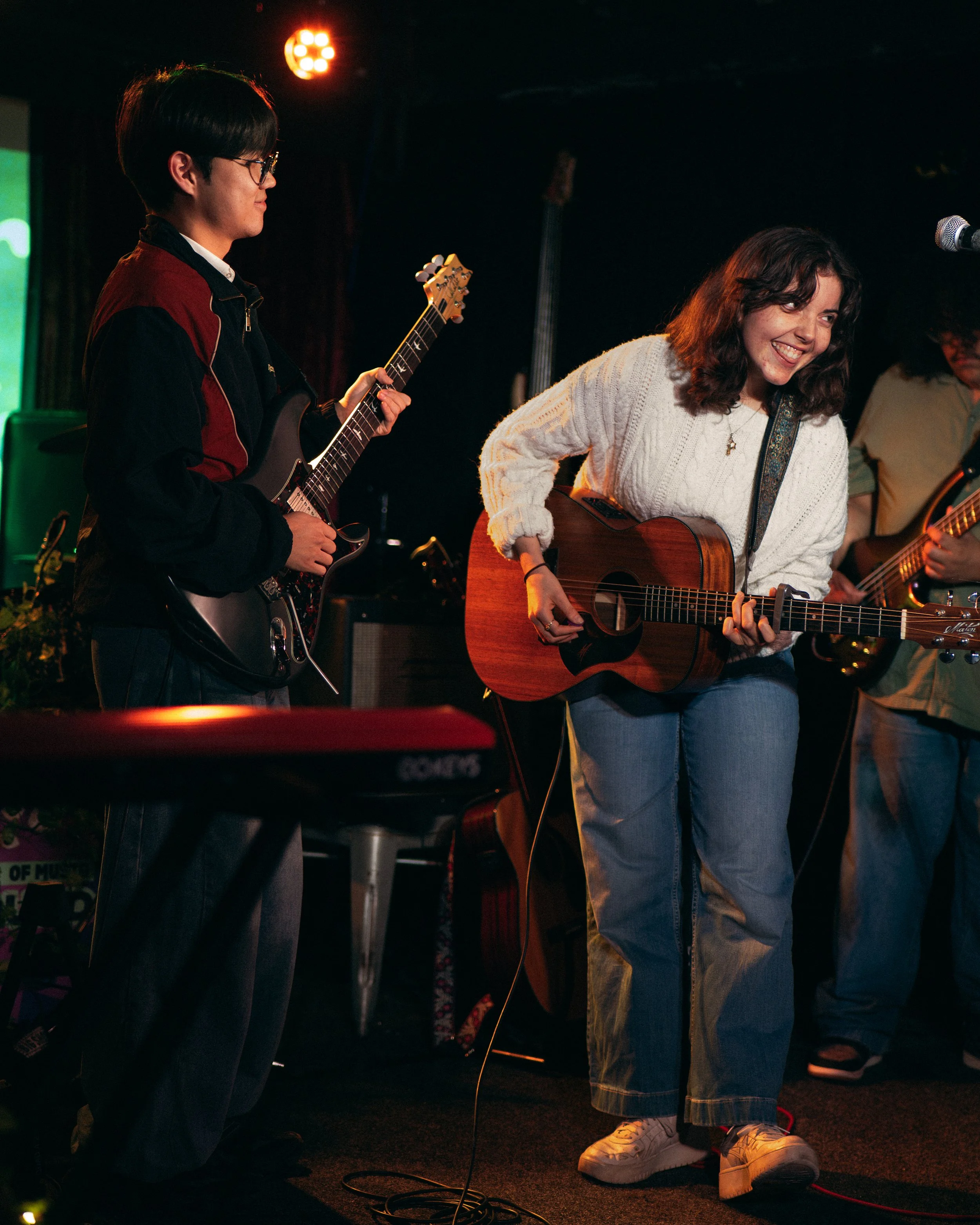 Young man and woman performing music together on stage at night. The young man is playing an electric guitar, and the young woman is playing an acoustic guitar and smiling.