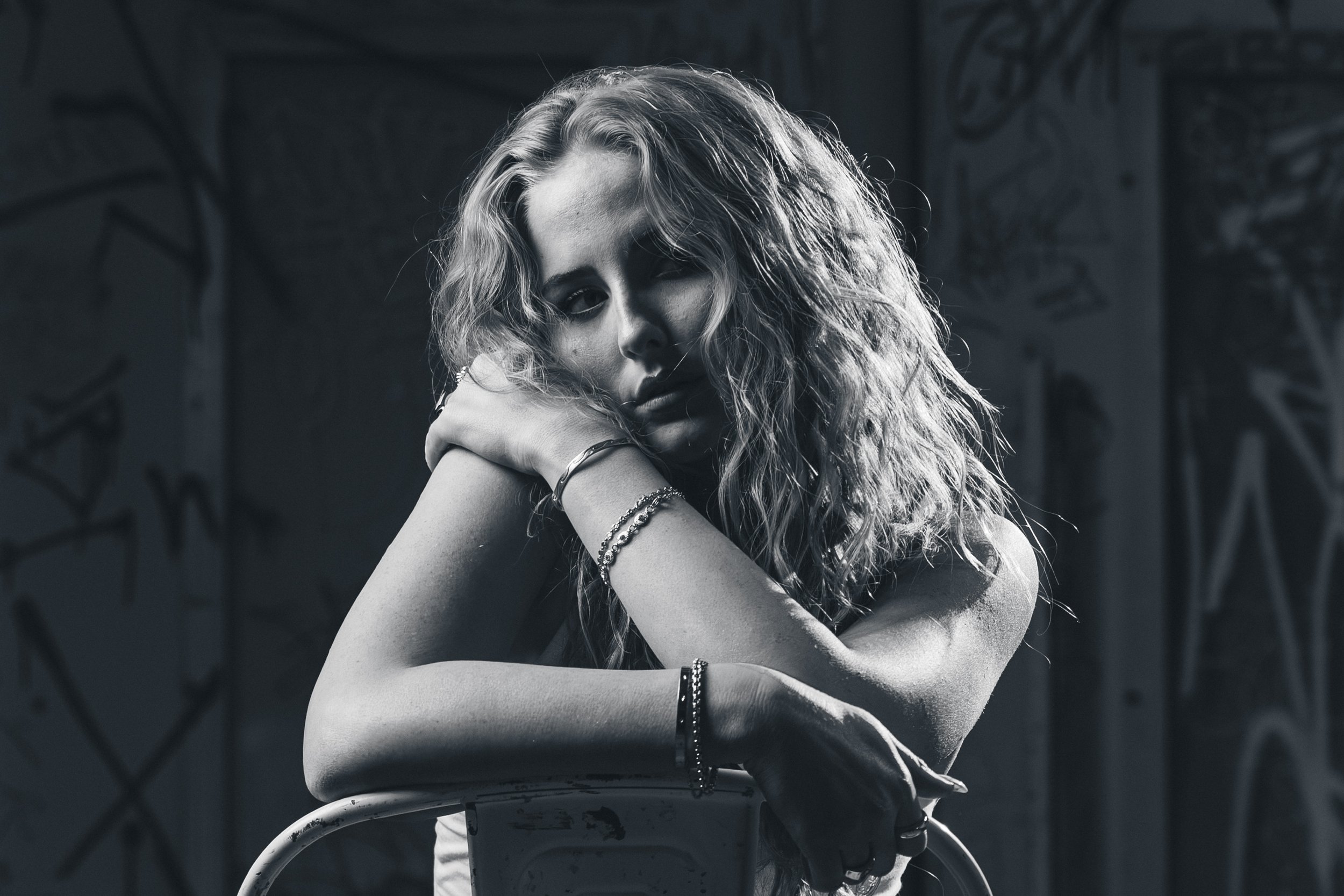A black and white photograph of a woman with wavy hair, resting her head on her hand, sitting on a bicycle in front of a graffiti-covered wall.