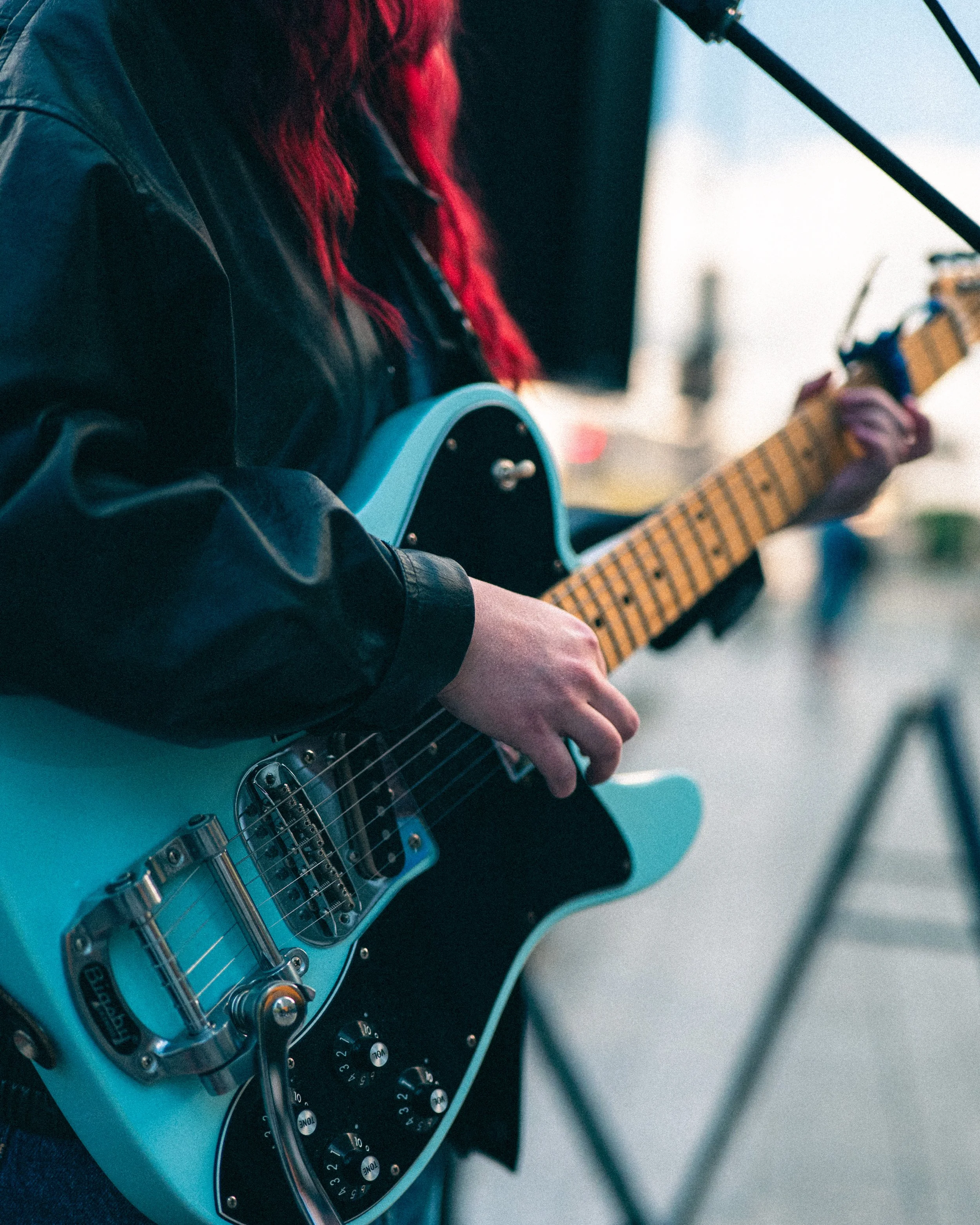 Close-up of a person playing an electric guitar with a blue body. The person is wearing a black jacket and has red hair. The guitar has various control knobs and a pickup selector switch, with a black pickguard. The person is holding the guitar neck 
