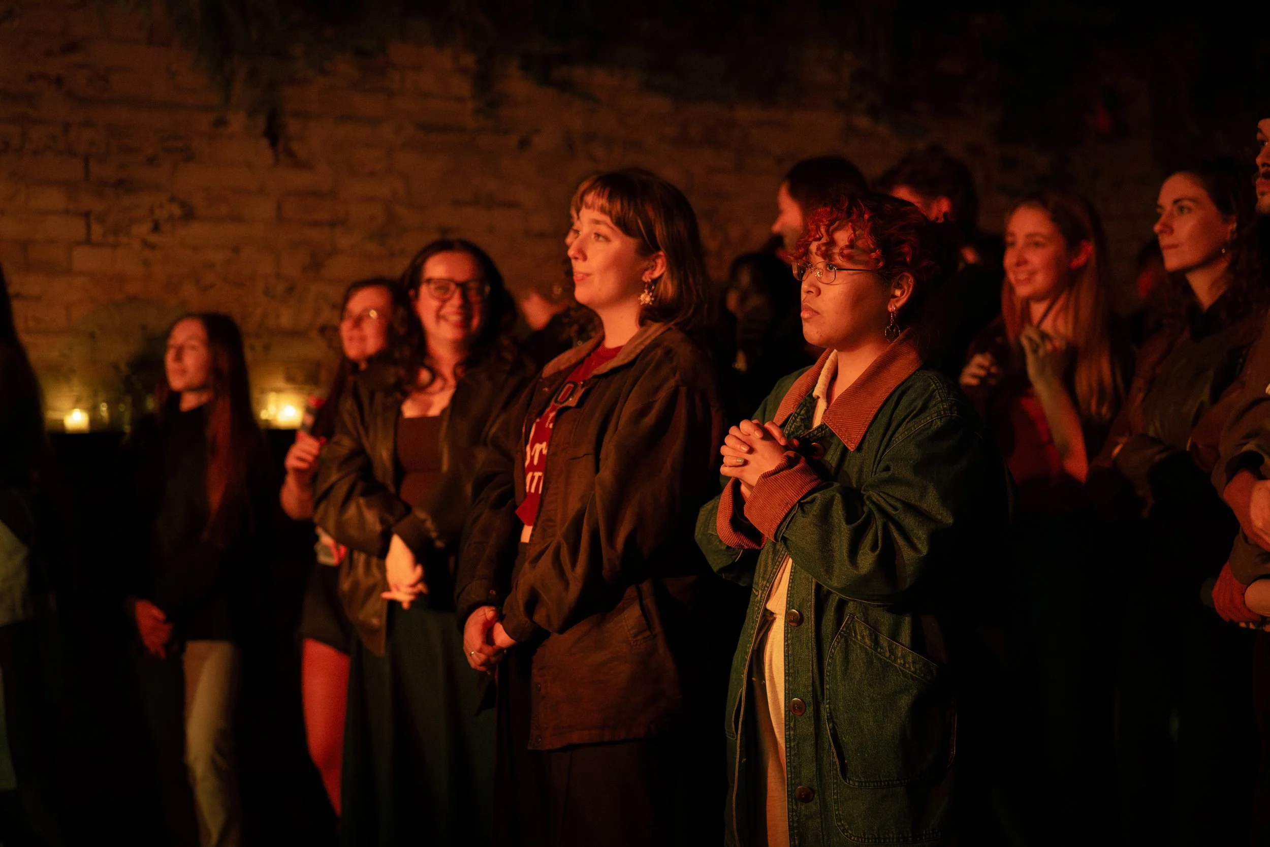 A group of people standing in a dimly lit space with a brick wall background, illuminated by warm, orange lighting, appearing engaged and attentive.