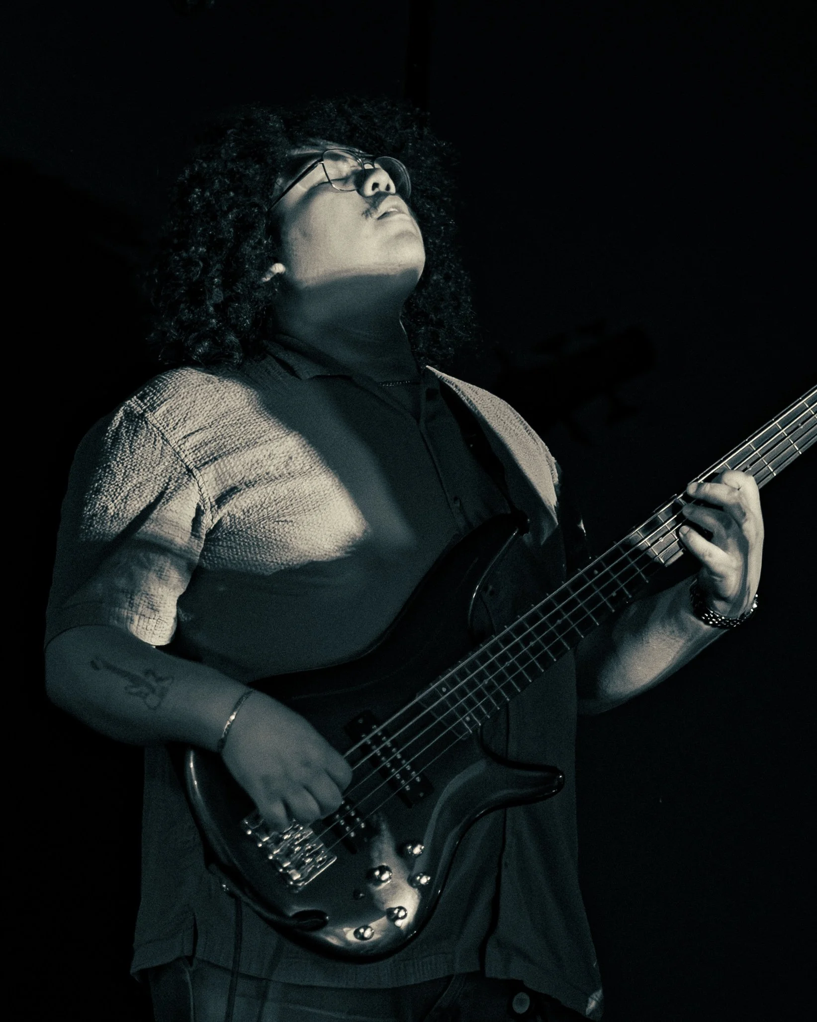 Person with curly hair and glasses playing an electric guitar in a dark setting.