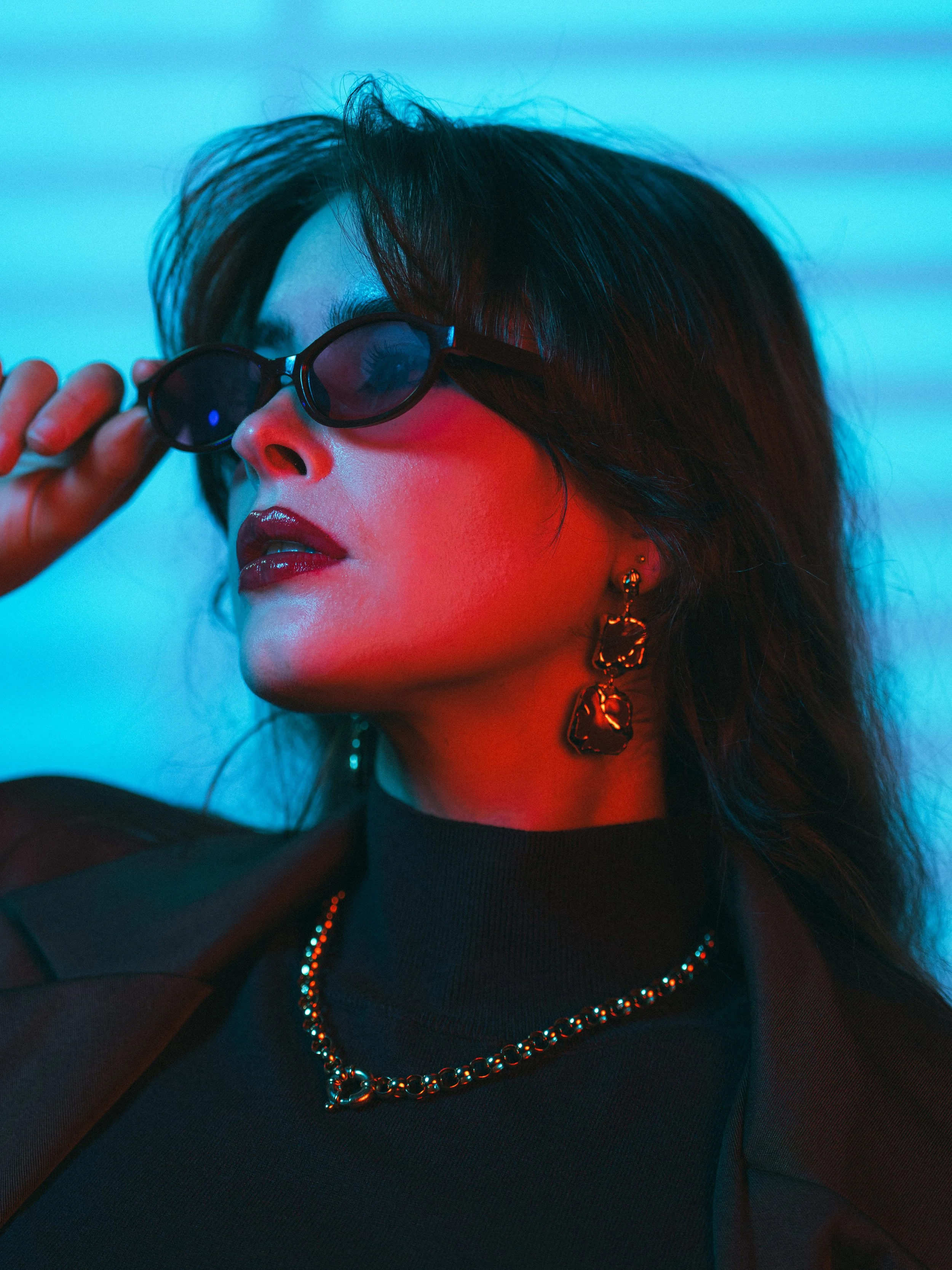 A woman with black hair wearing sunglasses, gold earrings, a black top, a gold chain necklace, and a black sheer jacket. The background is blue and red lighting.