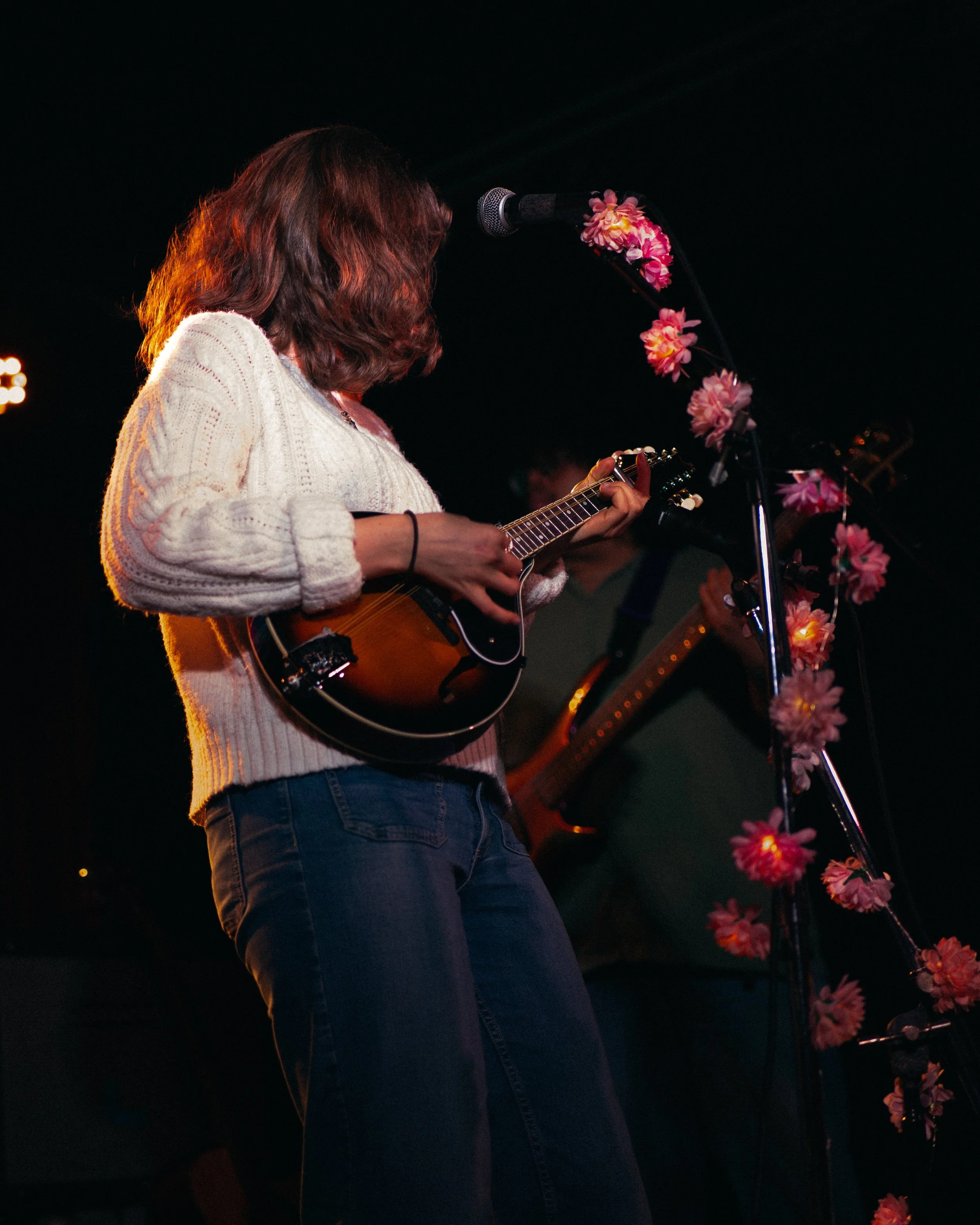 A woman with shoulder-length wavy hair wearing a light sweater, playing an acoustic guitar on stage decorated with pink flowers, in front of a microphone, with a dark background.