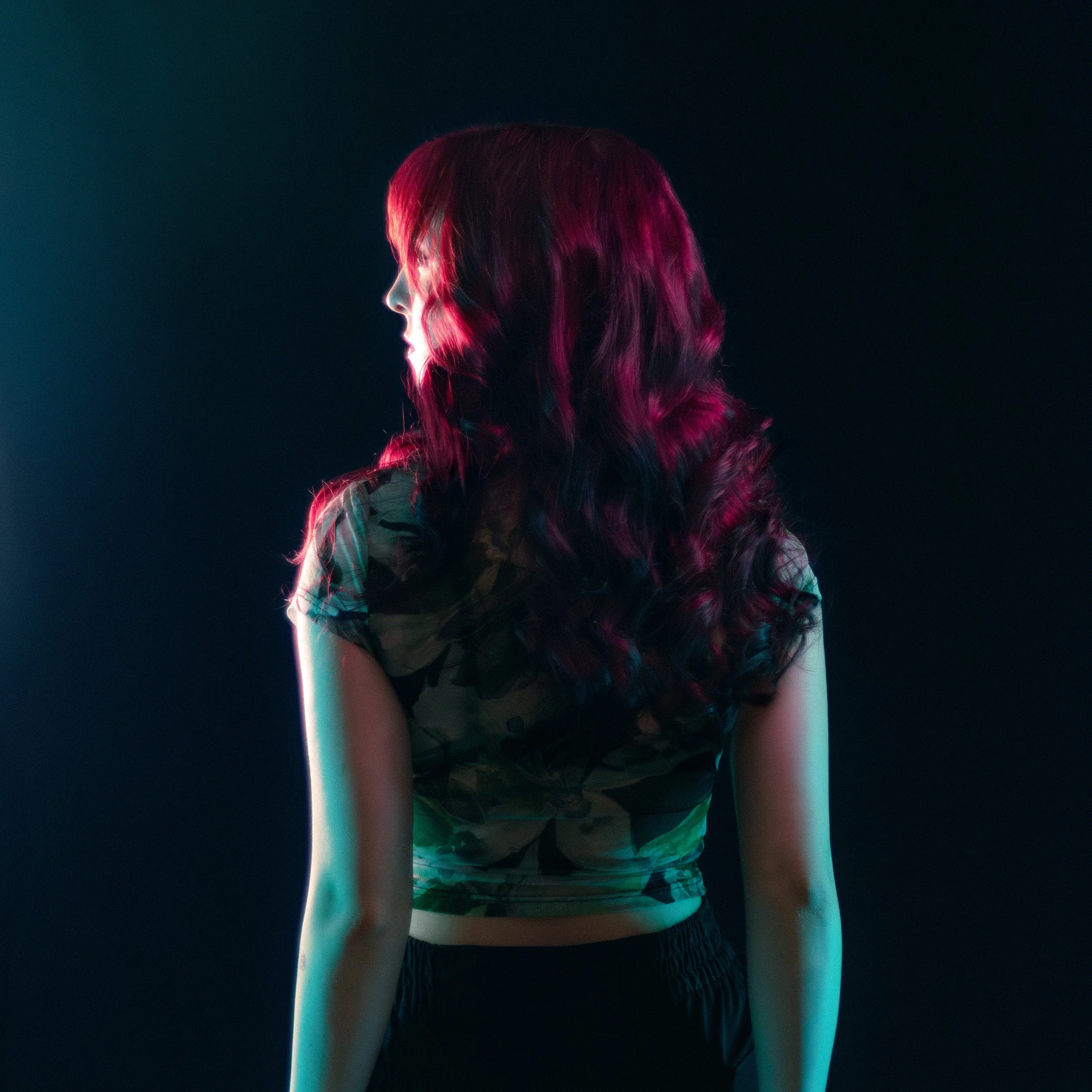 A person with shoulder-length wavy hair, dyed pink and purple, is seen from behind. They are wearing a camouflage-patterned shirt and black shorts. The background is dark and the scene is illuminated with colored lights, creating a vibrant atmosphere