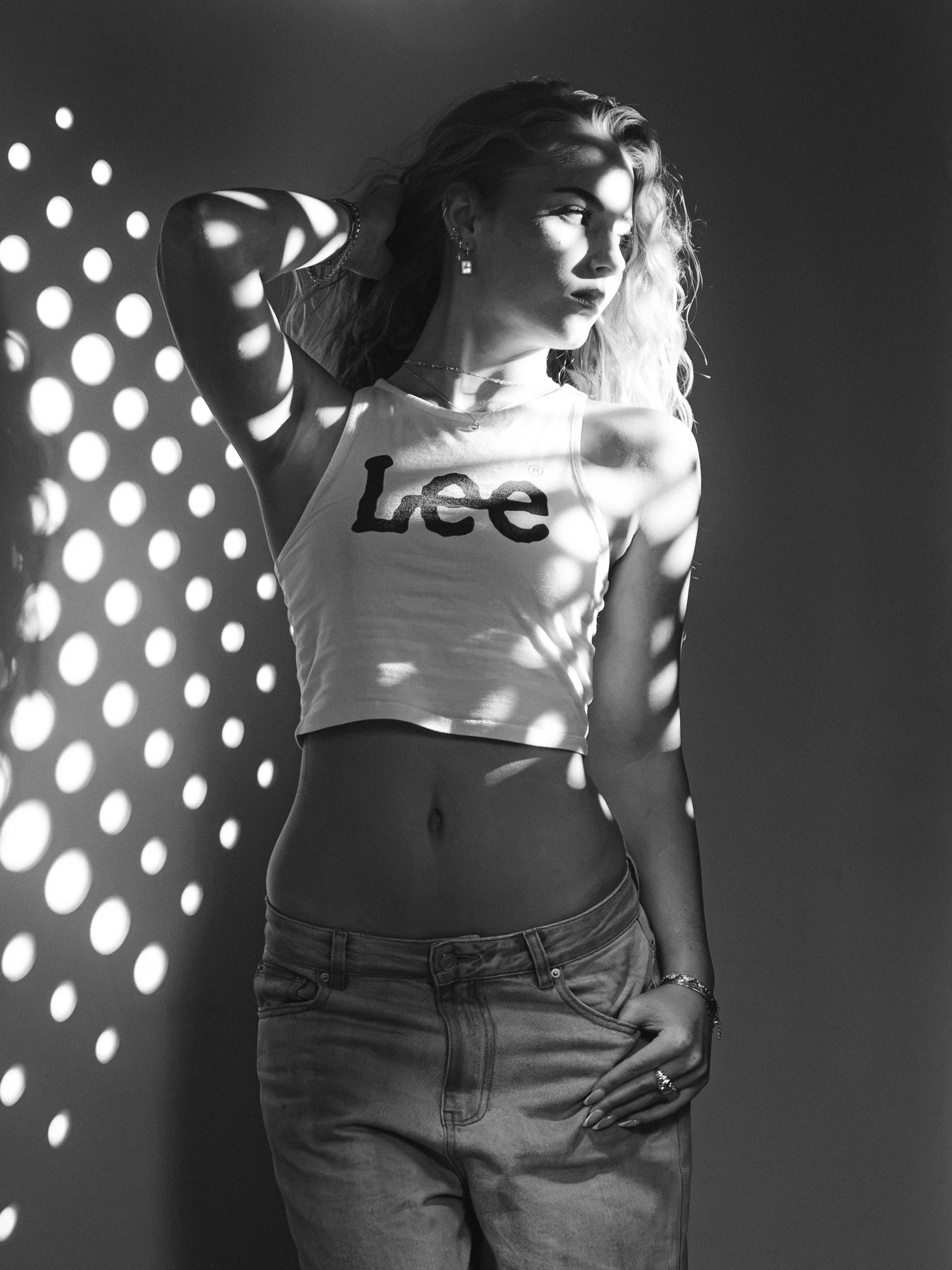 A woman with long, wavy hair stands in front of a dark background with light spots cast in a dot pattern. She is wearing a sleeveless top with the word 'Lee' printed on it and high-waisted jeans. She is posing with her right hand resting on her head 
