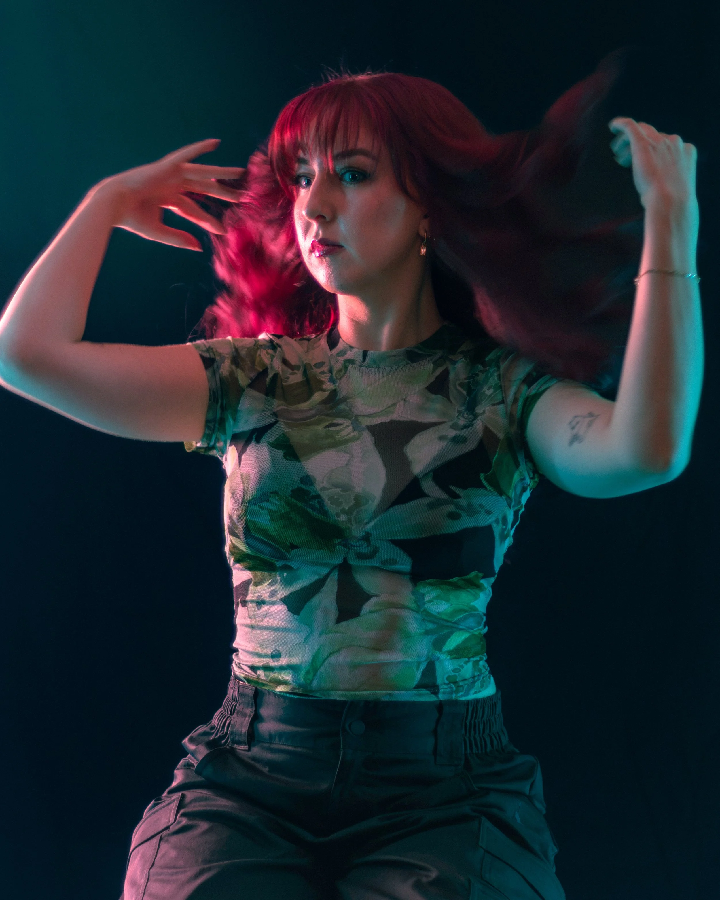 Woman with red hair and tattoos wearing camouflage t-shirt and black pants, with hands in hair, looking at camera against a dark background.