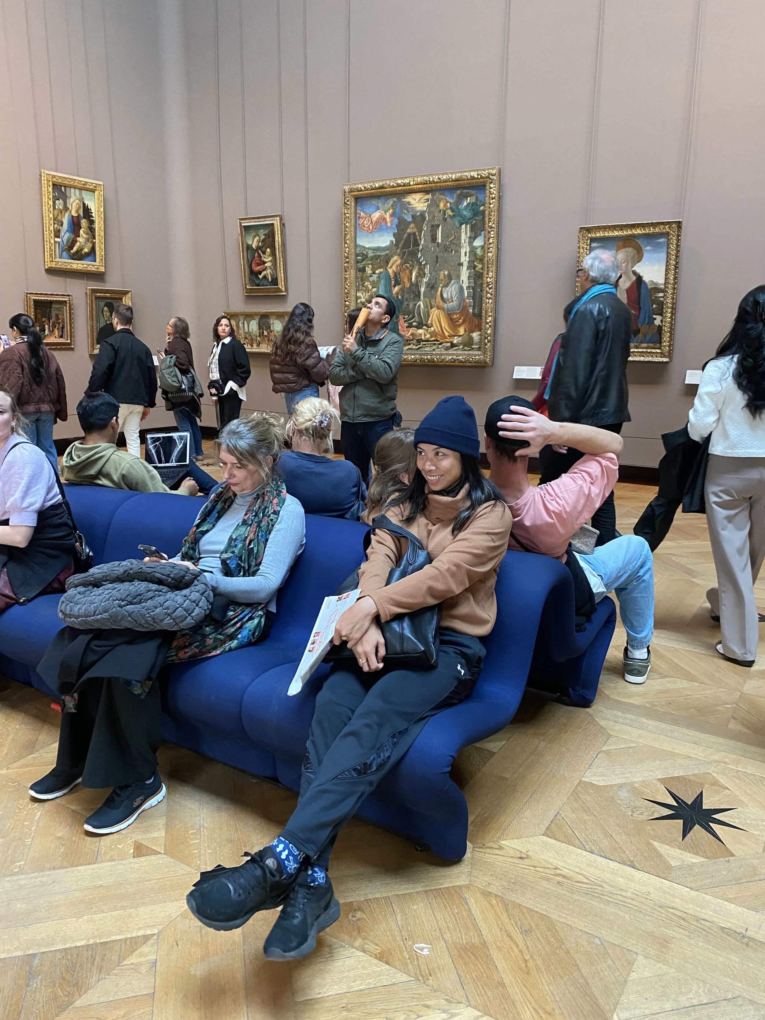 People at an art gallery viewing various paintings on the wall, some seated, some standing, with a man holding a book and a woman taking photos.