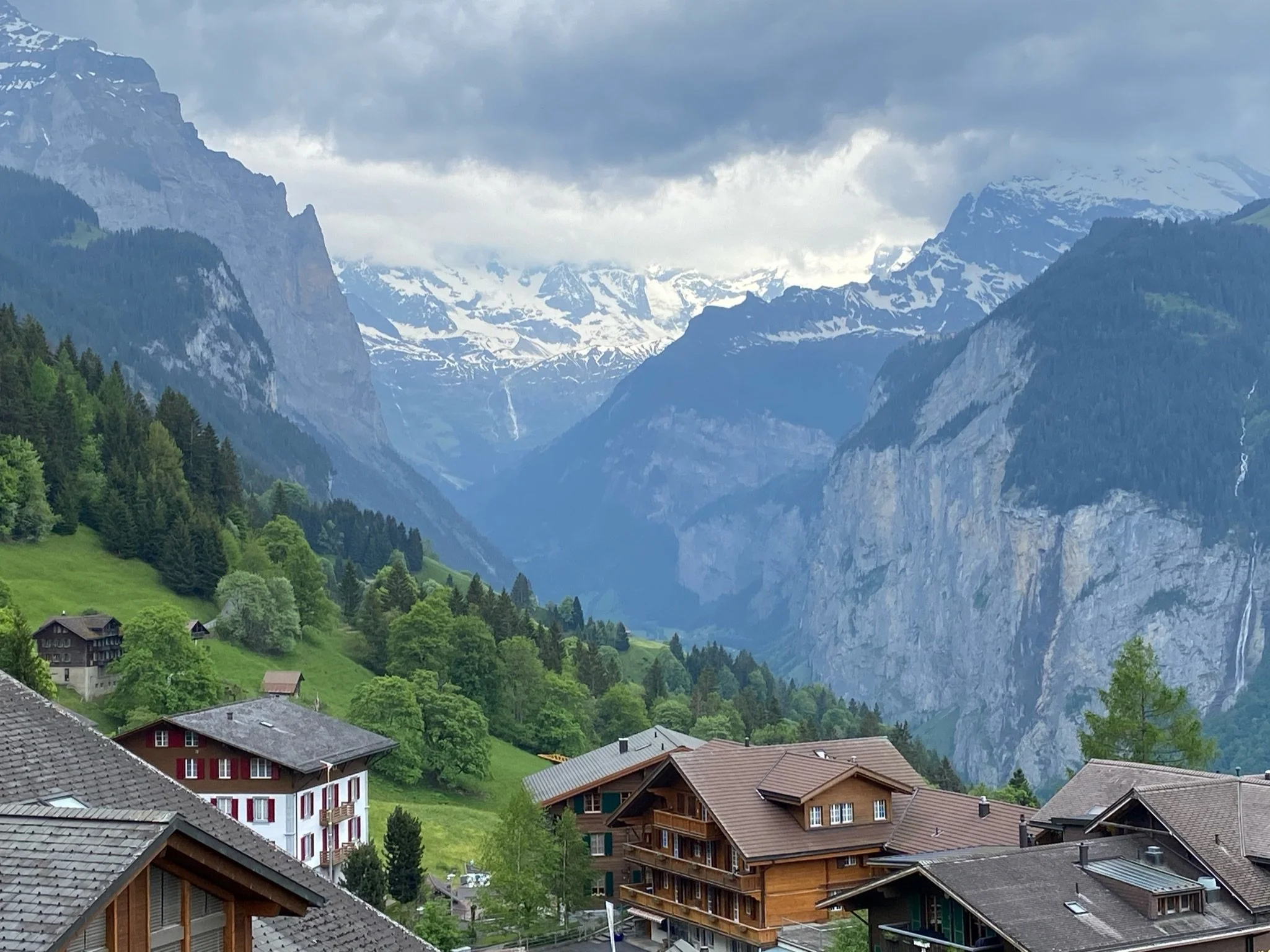 Mountain village with houses, surrounded by lush green trees and forested hills, mountain peaks with snow in the background under cloudy sky.