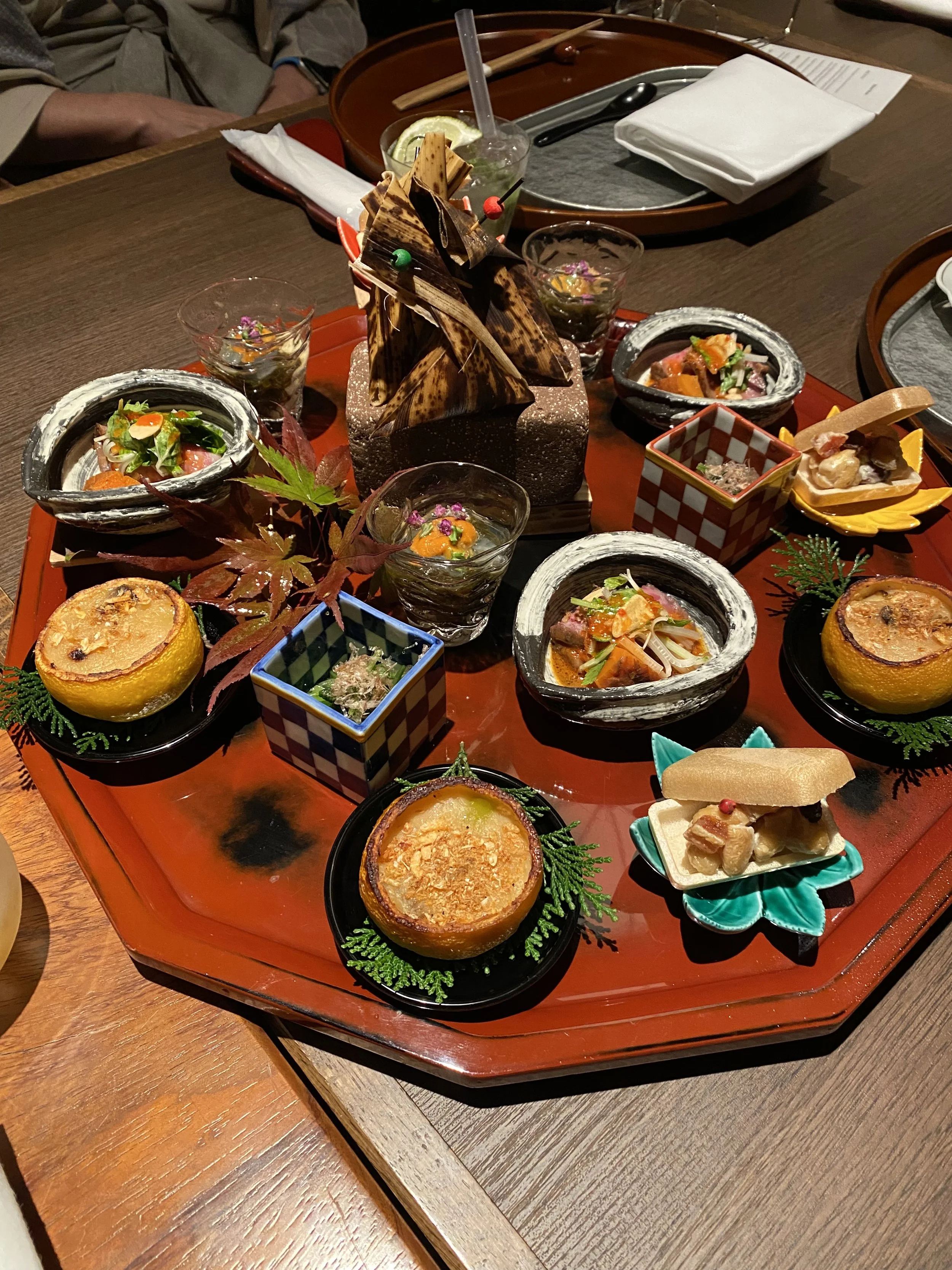A traditional Japanese food presentation on a wooden table, featuring various small dishes in bowls and cups, with leafy garnishes and a decorative arrangement of plated items.