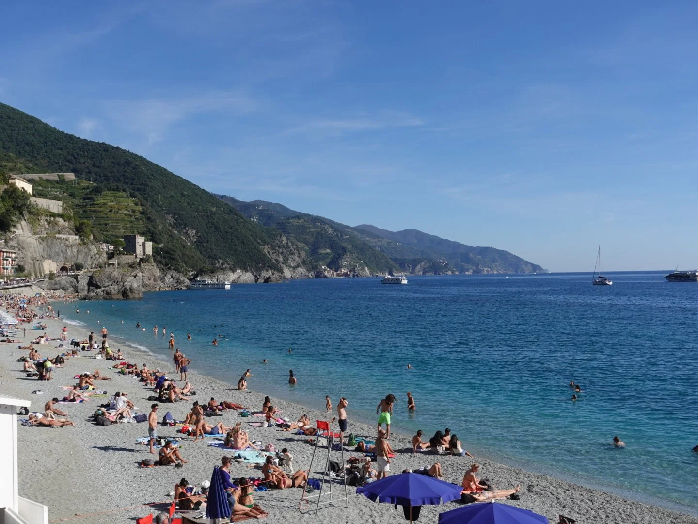 Crowded beach with people sunbathing and swimming, located on a rocky coastline with green hills and boats in the water, under a clear blue sky.