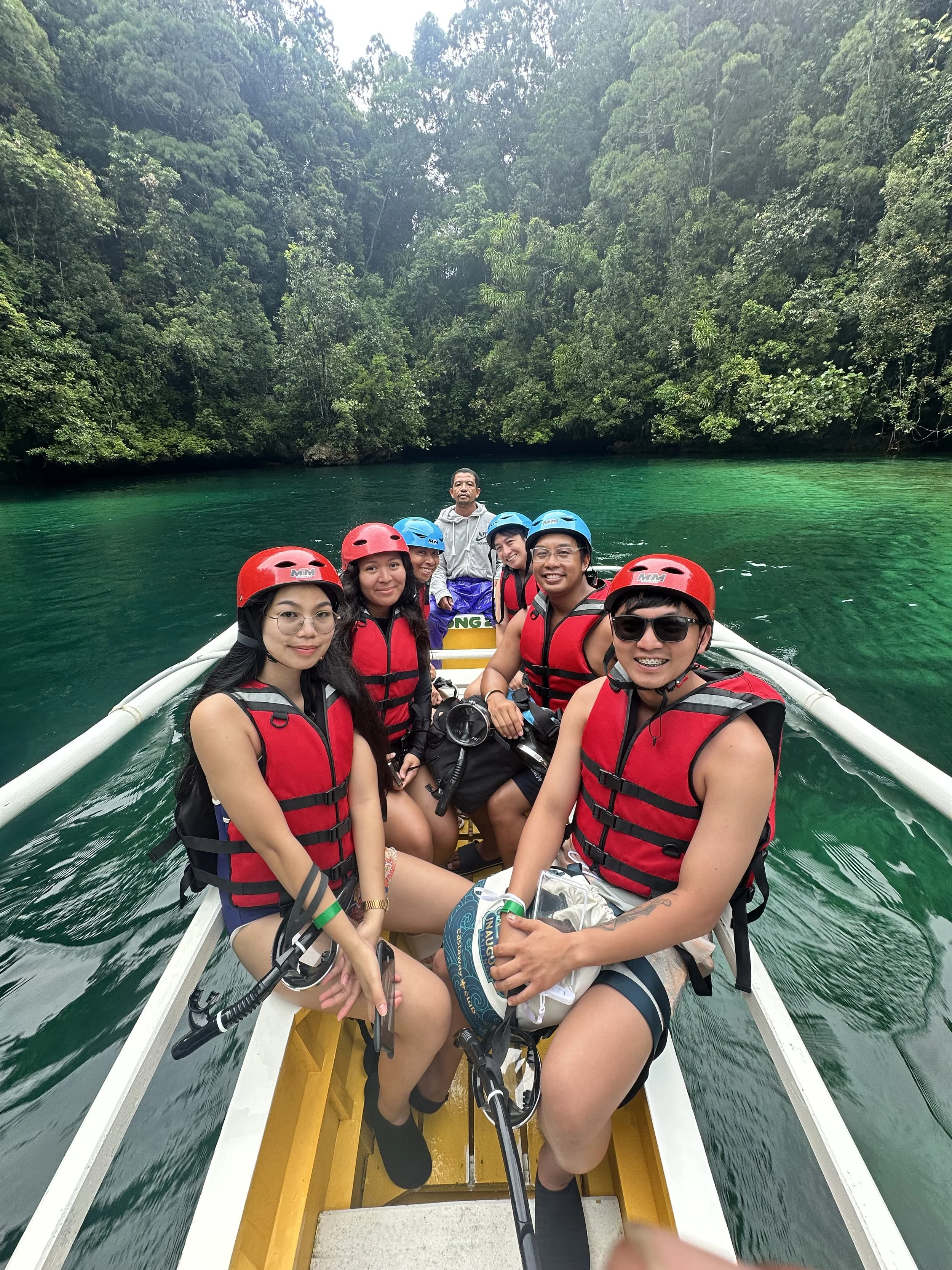 Group of people on a boat in a lush green river with dense trees in the background, wearing life jackets and helmets, smiling for the photo.