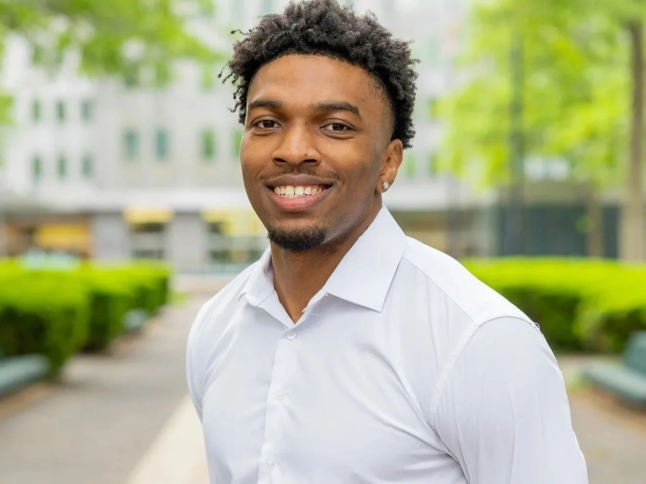 A young man with curly hair, earrings, and a white shirt standing outdoors in a park with green trees and bushes in the background, smiling at the camera.