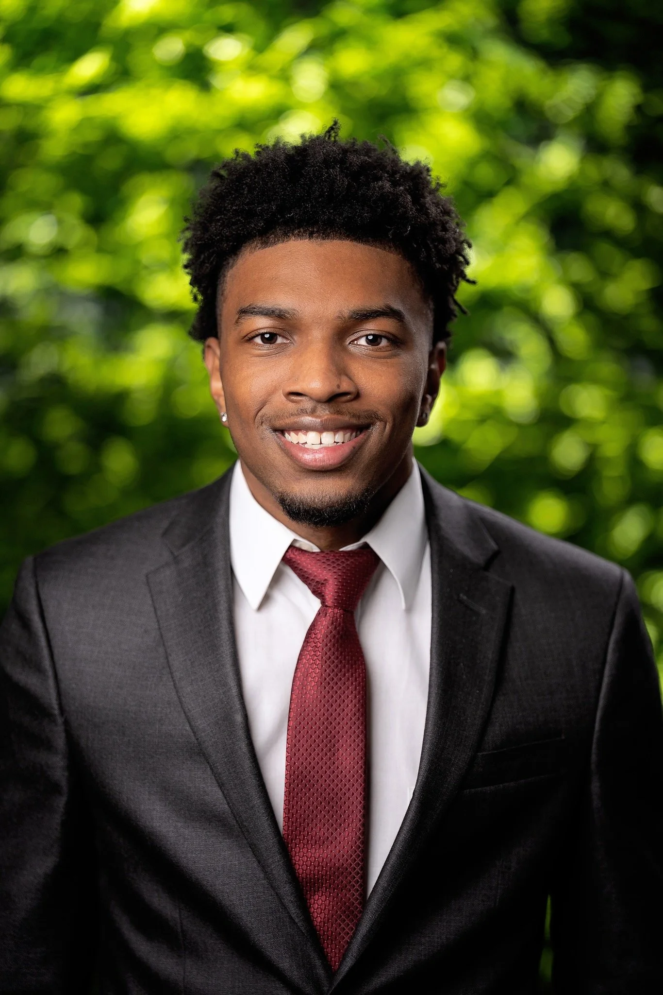 A young man in a dark suit, white shirt, and maroon tie, smiling with a blurred green leafy background.