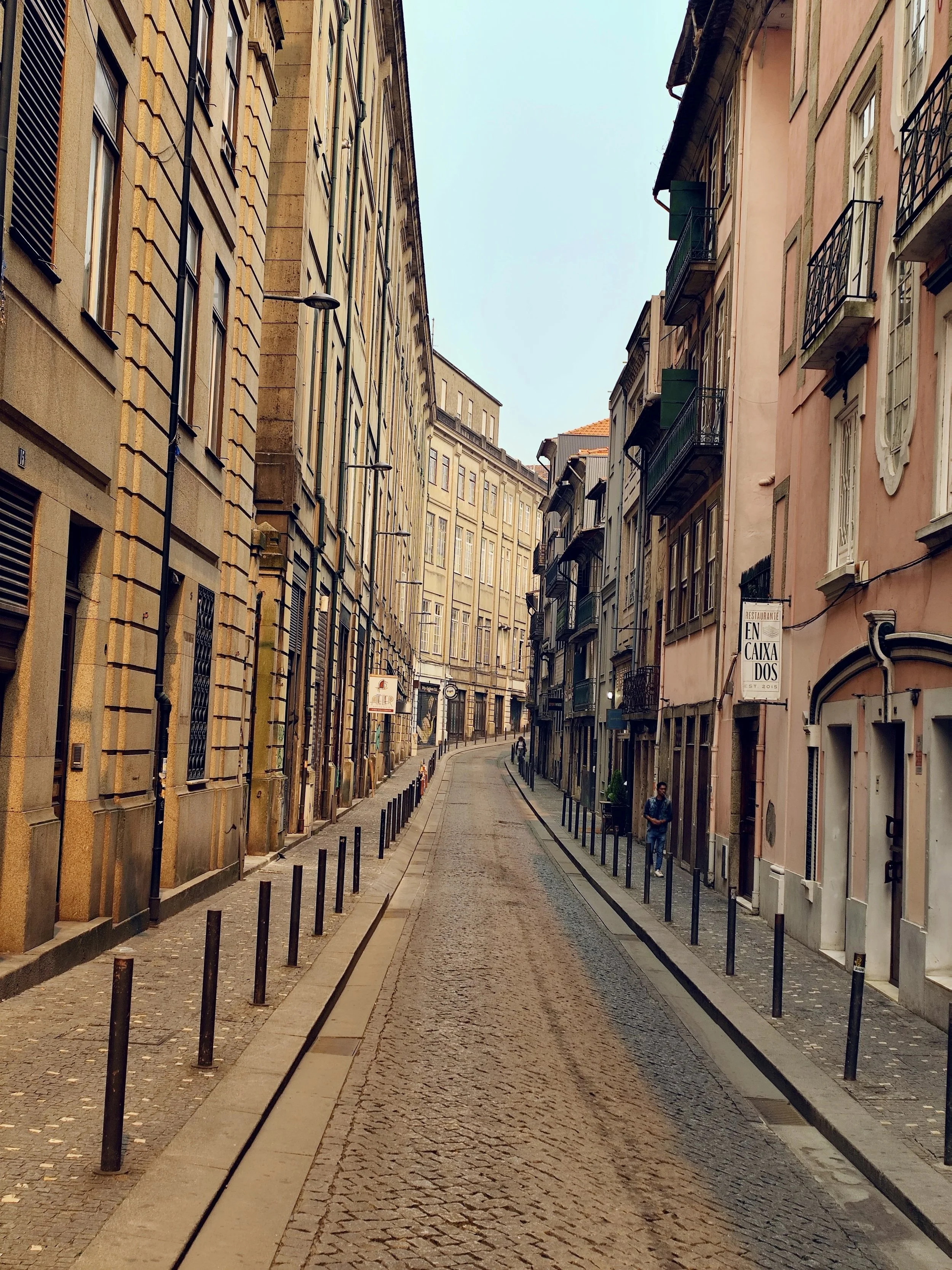 a narrow cobblestone street lined with tall old buildings, windows and small balconies, with a few pedestrians walking.