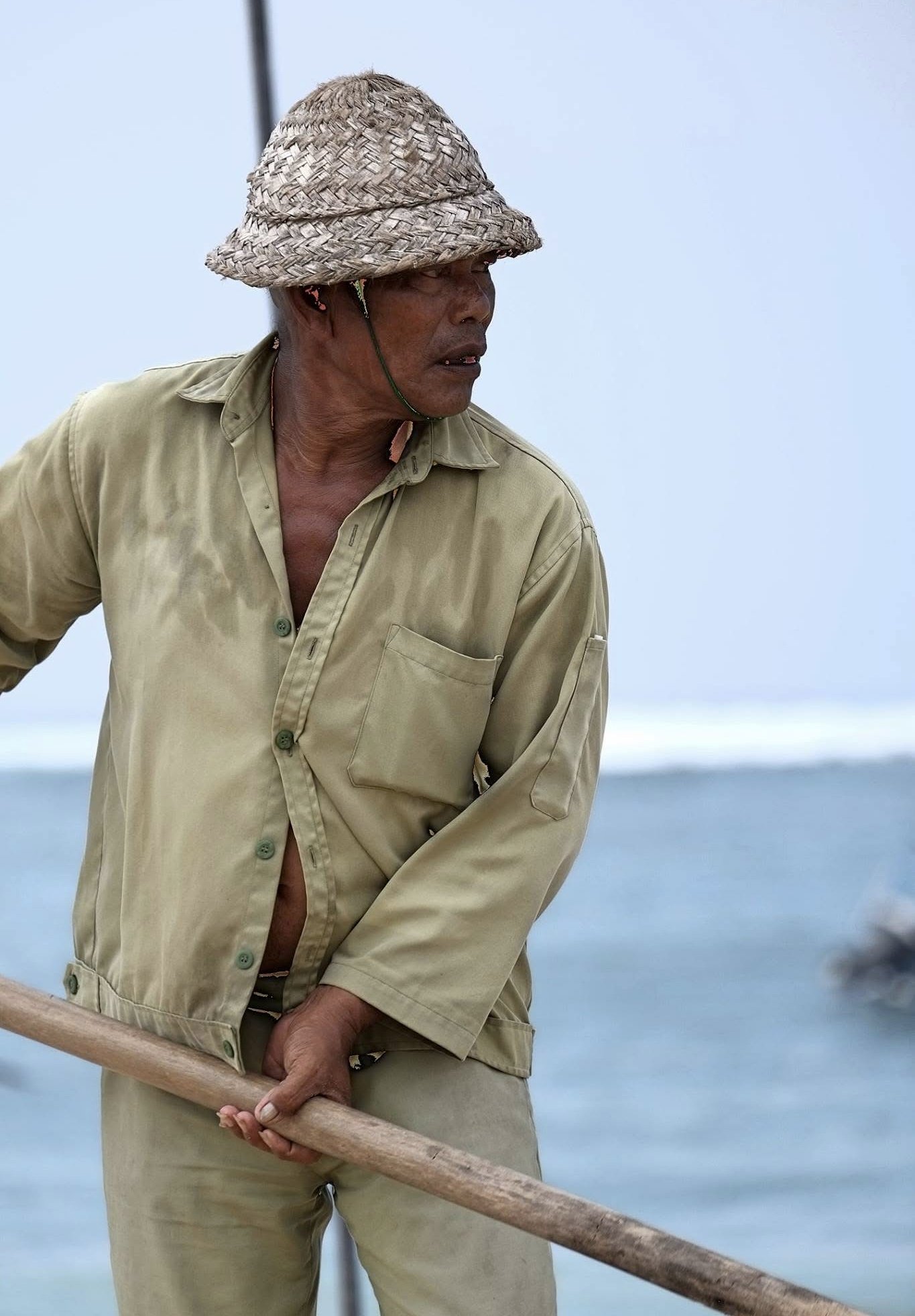 a closer view of a man in a hat and light clothing holding a long wooden tool on a beach in bali.