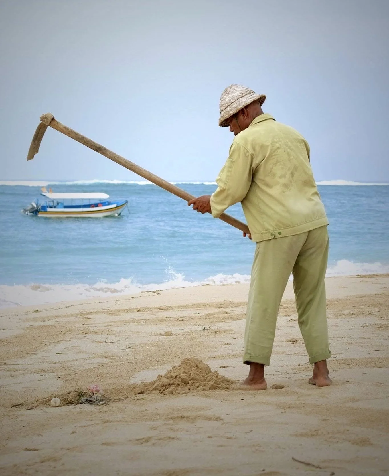a barefoot man in a hat working on a sandy beach in bali, with a boat on the sea behind him.