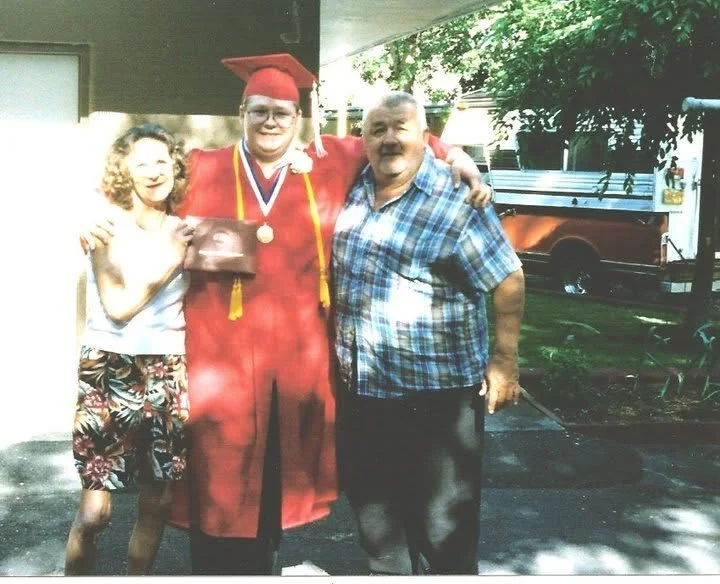 A young man in a red graduation cap and gown holding a diploma, standing between an older man in a plaid shirt and a young girl in a white shirt and patterned shorts, outdoors with trees and a vehicle in the background.