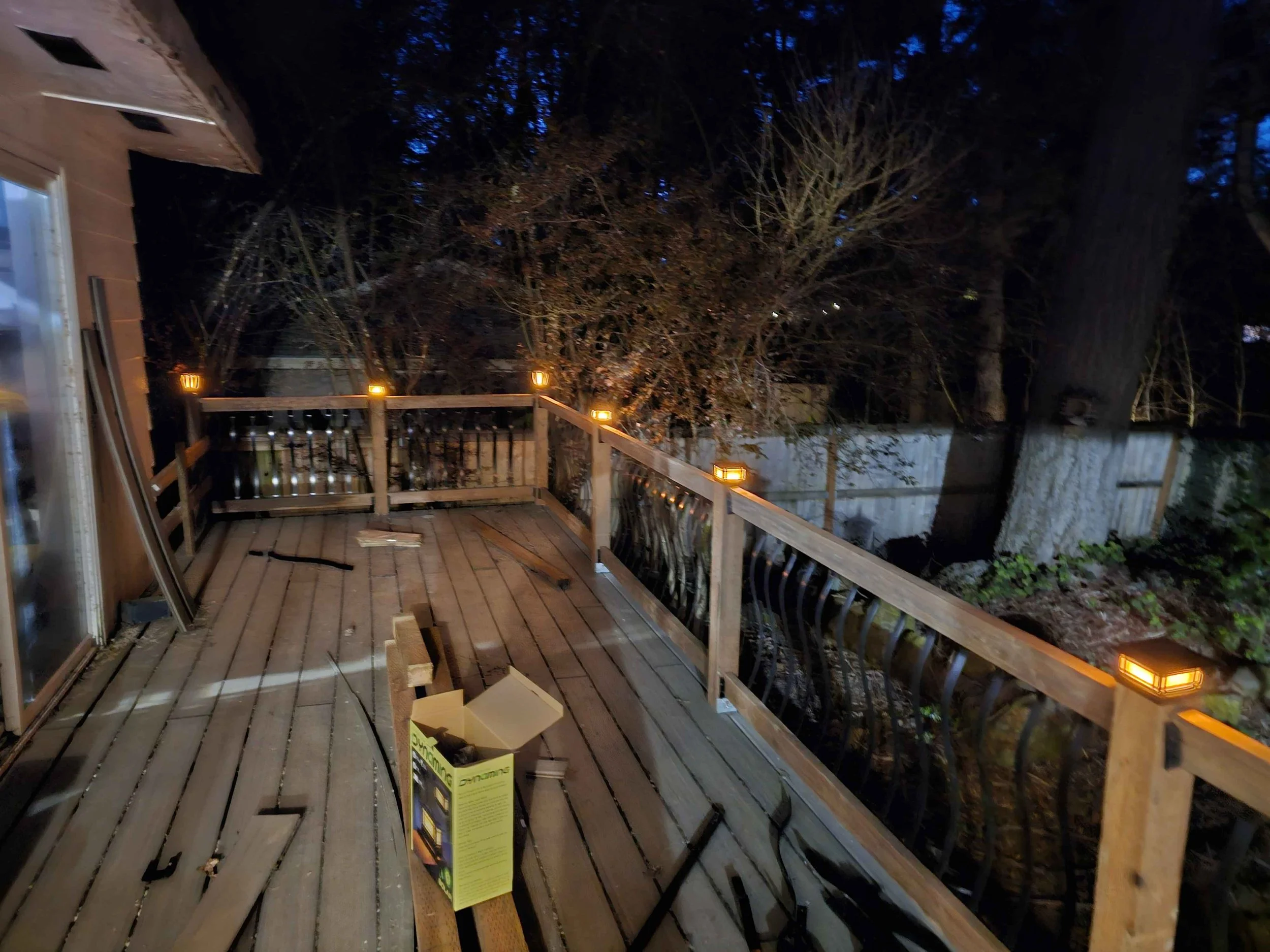 A wooden deck with a railing and decorative lantern lights, surrounded by trees at night. There is a yellow and black product box on the deck, and construction materials are nearby.