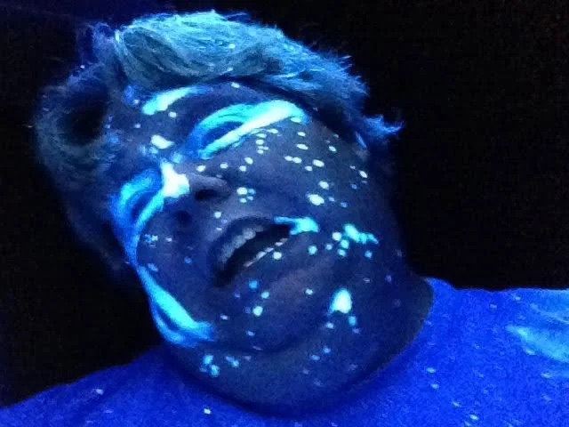 Person smiling and laughing with face illuminated by glowing blue and white lights, possibly under a blacklight or glow-in-the-dark effects.