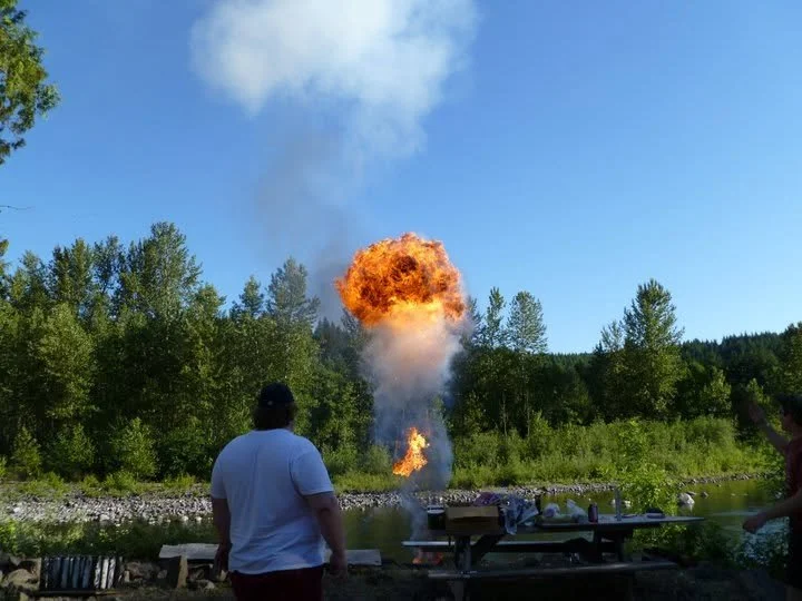 A large explosion or fireball erupts from a protest site with a blue sky and green trees in the background. Two people are observing the scene, with one person in the foreground watching the explosion.