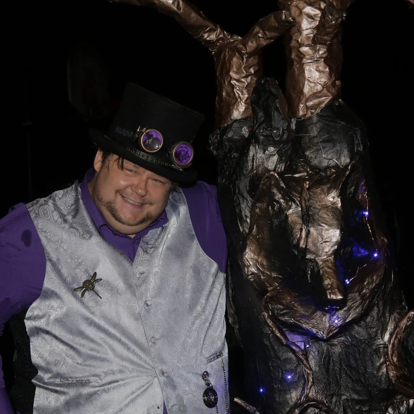 A man wearing a purple shirt, silver vest, and a black hat decorated with purple and black buttons is smiling. He has a beard and is standing next to a Halloween or creepy decoration that looks like a black and brown twisted figure with lights attached.
