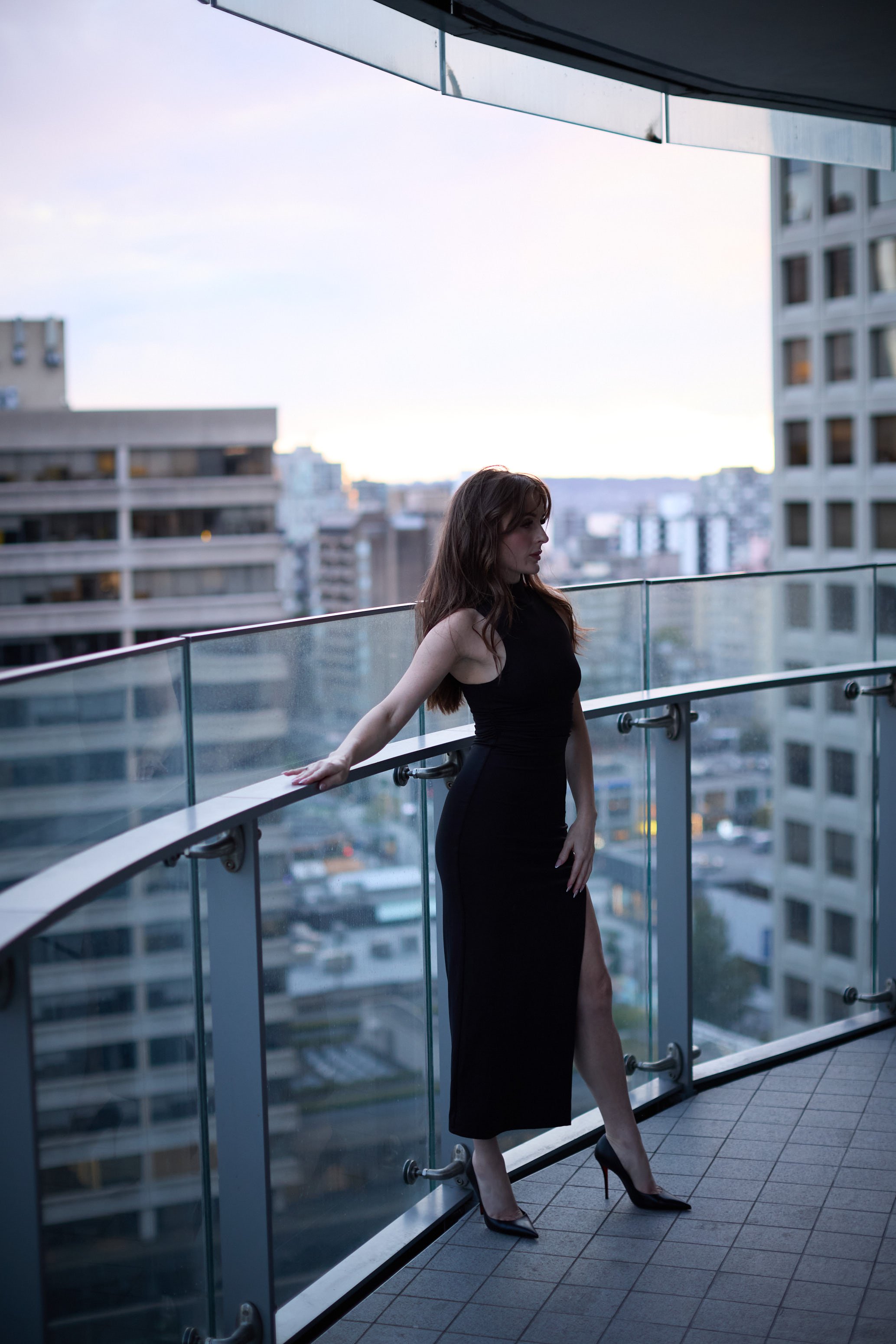 A woman with long red hair wearing a black dress and high heels standing on a balcony with glass railing, overlooking a cityscape of tall buildings during dusk or early evening.