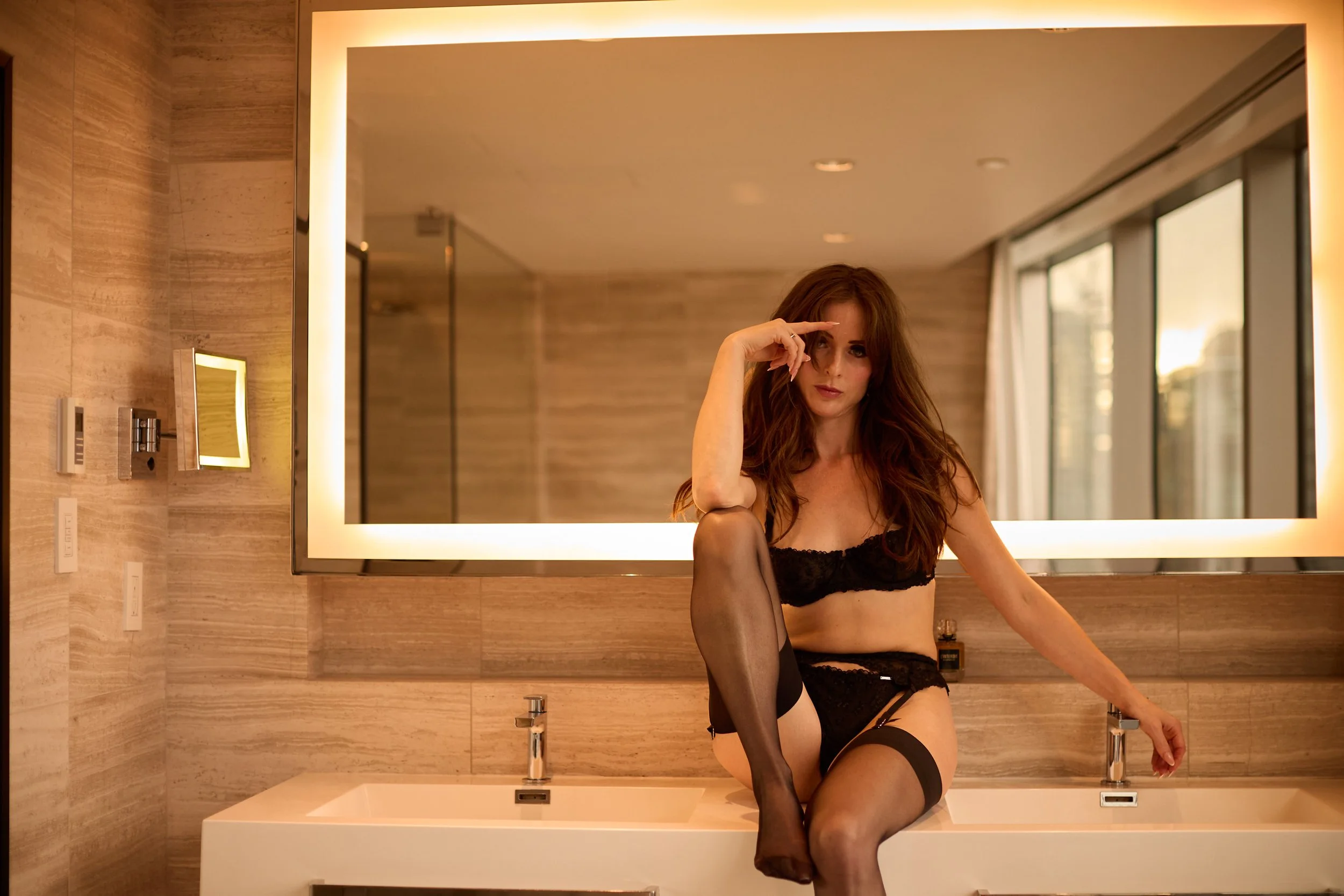 A woman with long red hair in black lingerie and stockings sitting on a bathroom sink in front of a large illuminated mirror.