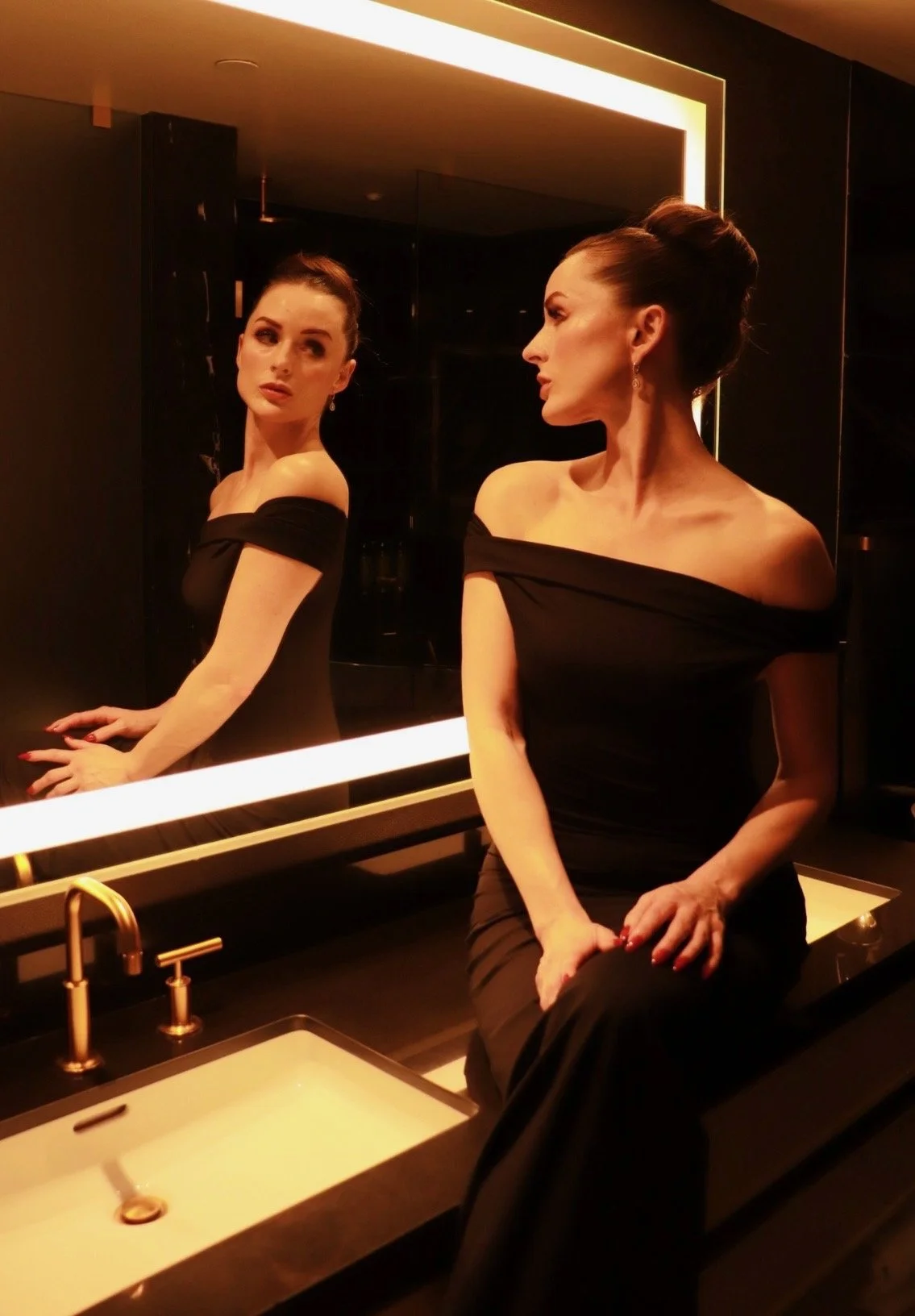 A woman in a black off-the-shoulder dress sitting by a sink in front of a mirror, looking at her reflection. The room has warm lighting and a dark background.