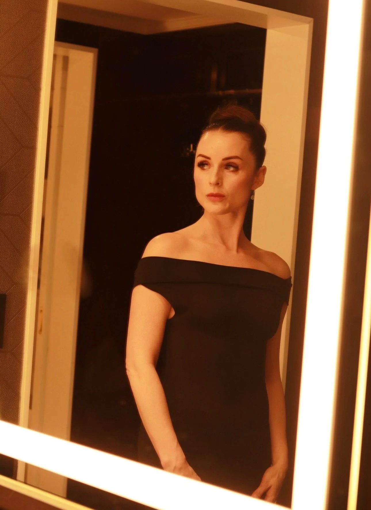 A woman with dark hair styled in an elegant updo, wearing a black off-the-shoulder dress, looking into a mirror with warm lighting.