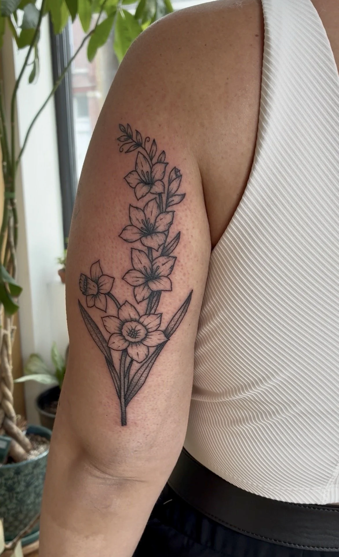 A person's upper arm with a black outline tattoo of a floral design featuring various flowers and leaves, with a background of indoor plants and windows.