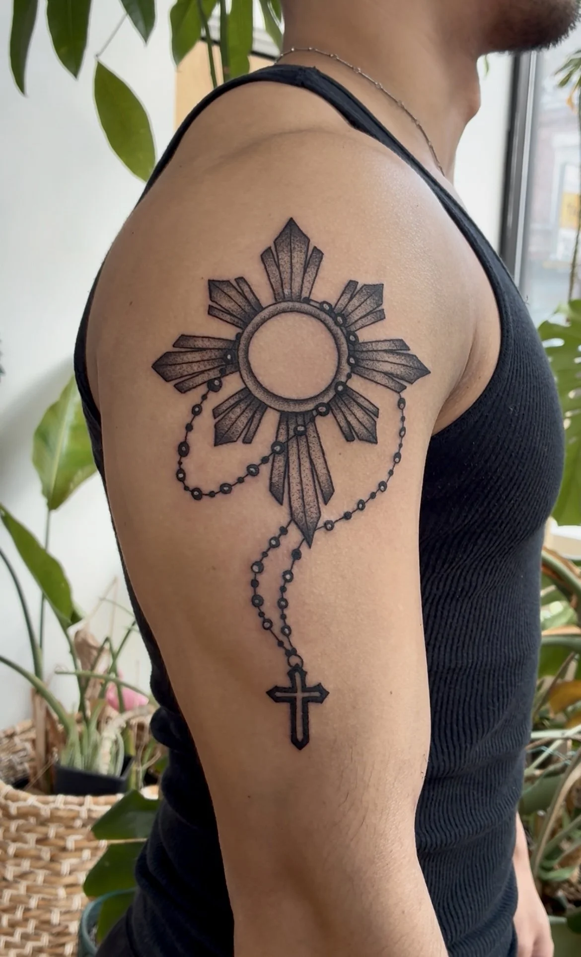 Tattoo of a sunburst design with a circle at the center on a person's upper arm, connected to a chain of beads and a cross hanging at the bottom.